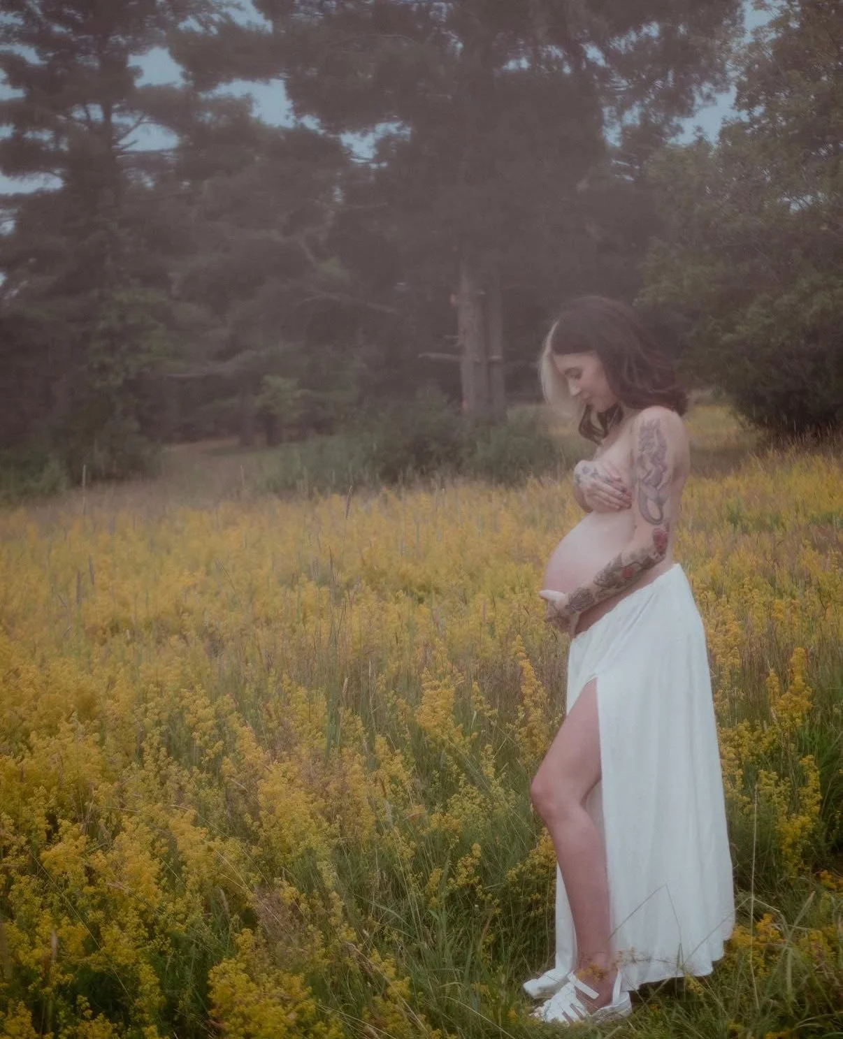 A pregnant woman with tattoos on her arms standing in a field of yellow flowers, wearing a white skirt and white sneakers, with her hands covering her breasts and looking down at her belly.