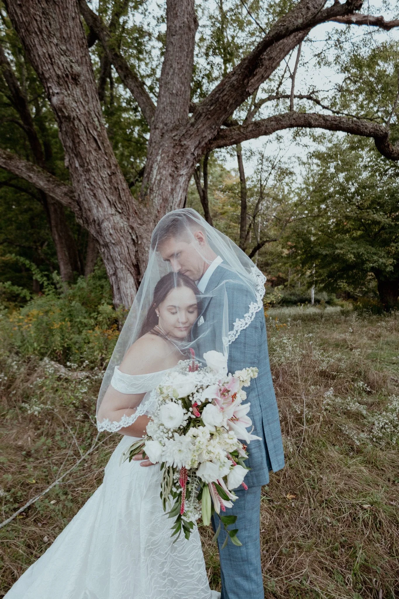 A bride and groom embrace under a veil in a natural outdoor setting with trees and grass, with the bride holding a large bouquet of white and pink flowers.