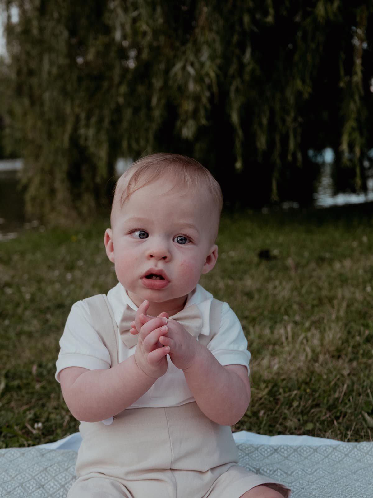 A young child with light skin and short light brown hair, dressed in a white shirt with a bow tie and beige overalls, sitting on a blanket outdoors near a body of water with trees in the background, with hands clasped together and a curious expressio