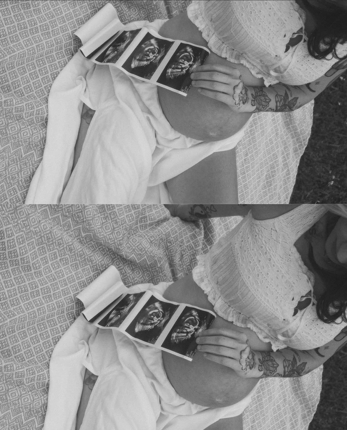 A pregnant woman lying on a patterned bed, holding ultrasound images near her belly, with tattoos on her arm, wearing a white knit top and skirt.
