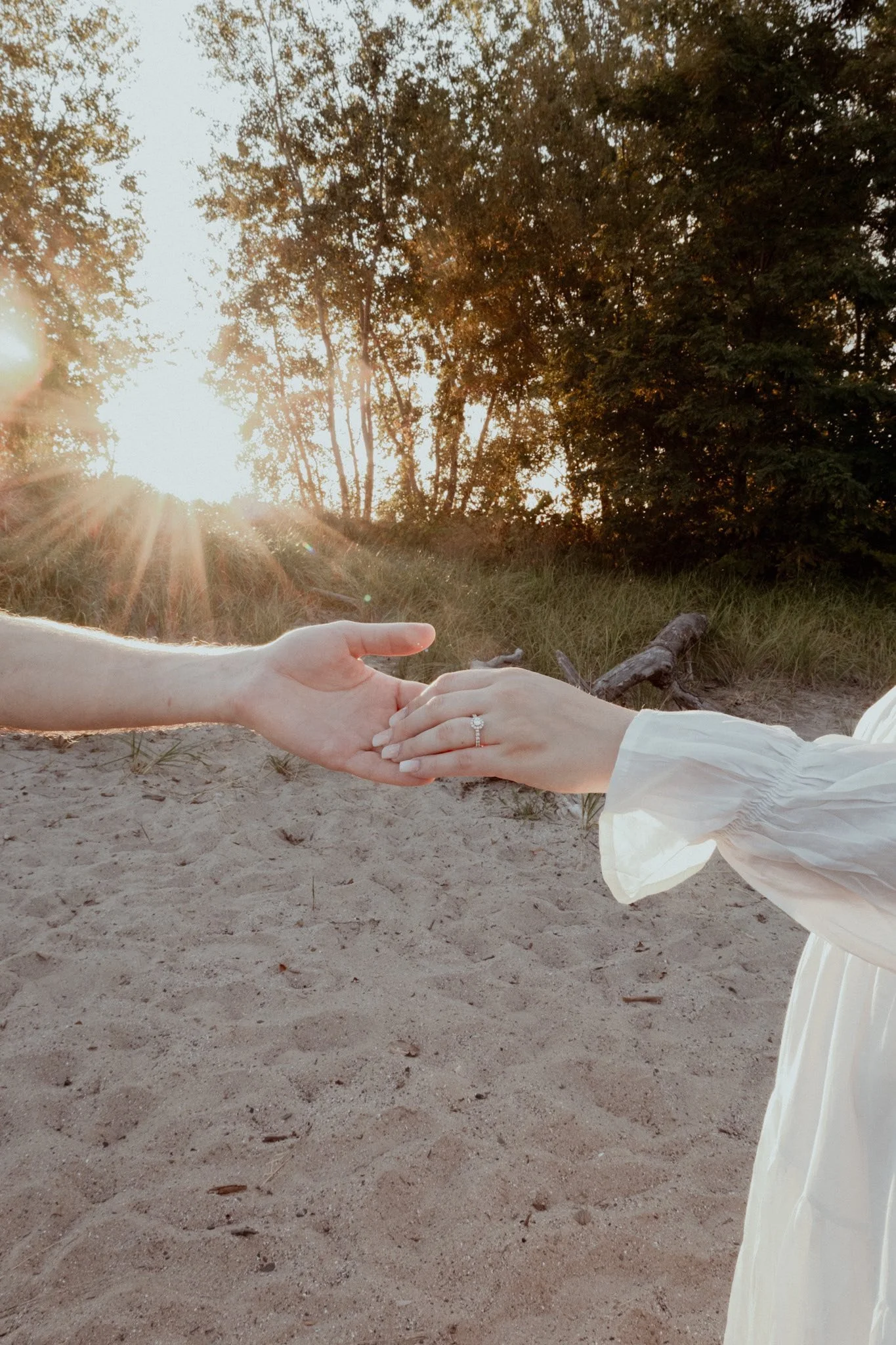 A person in a white dress holding hands with someone else, with a sunset in the background at the beach.