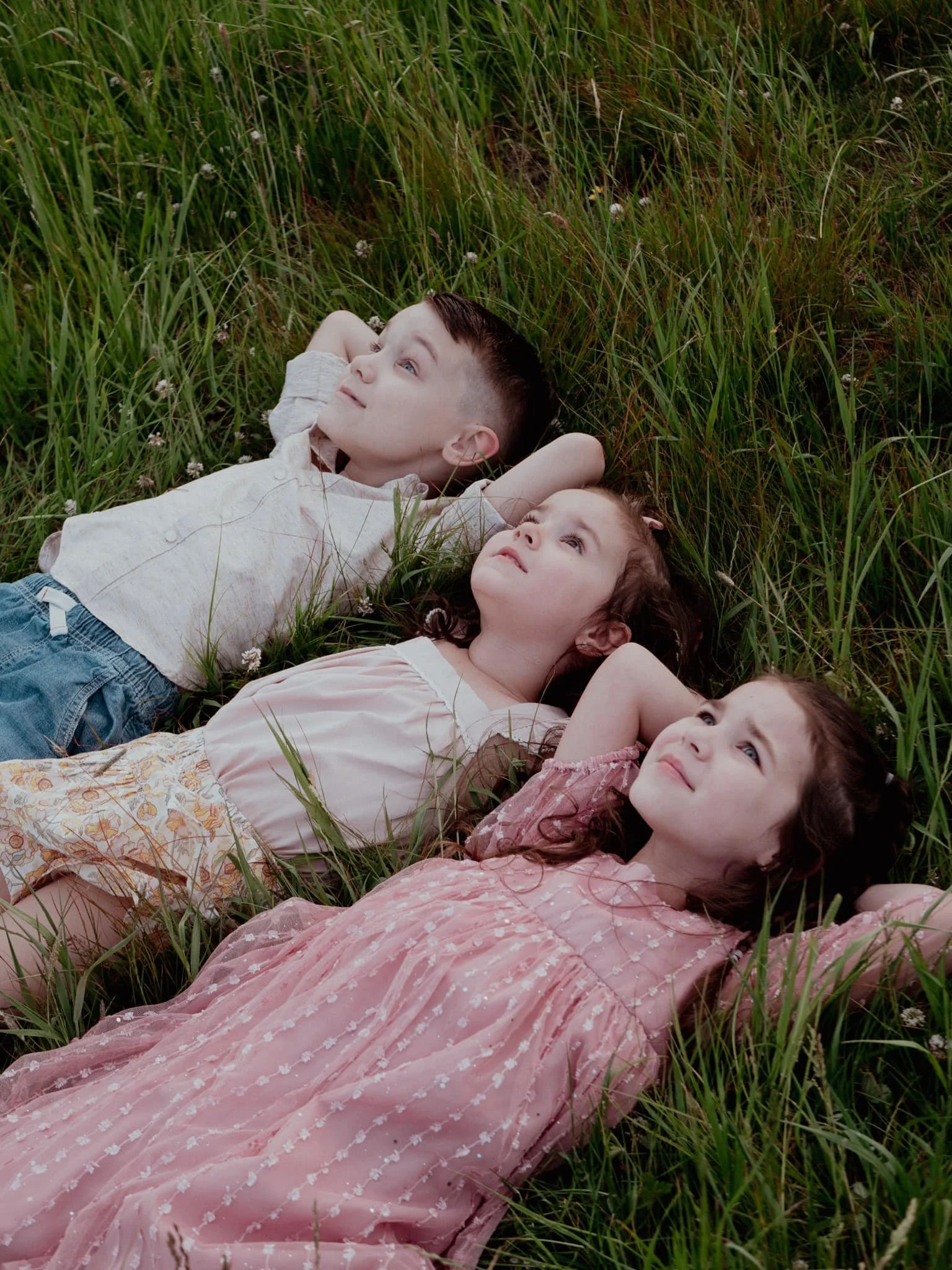 Three children lying on grass, gazing upward, outdoors in a grassy field.