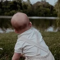 Young child with a shaved head sitting on grass near a body of water, facing away from the camera.
