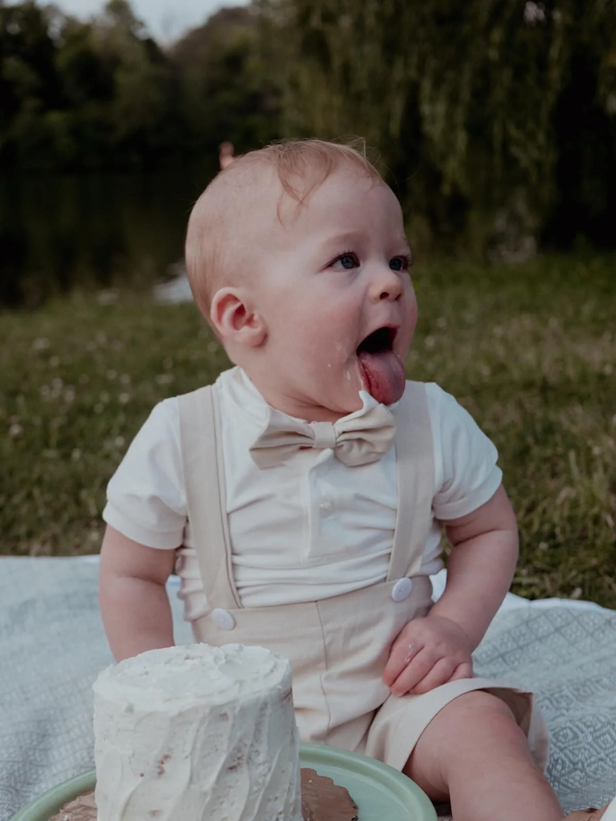 A baby boy with fair skin and light hair, wearing a white shirt with a bow tie and beige suspenders, sitting on a blanket outdoors near a body of water with trees in the background. The baby is sticking out his tongue and appears to be excited or pla