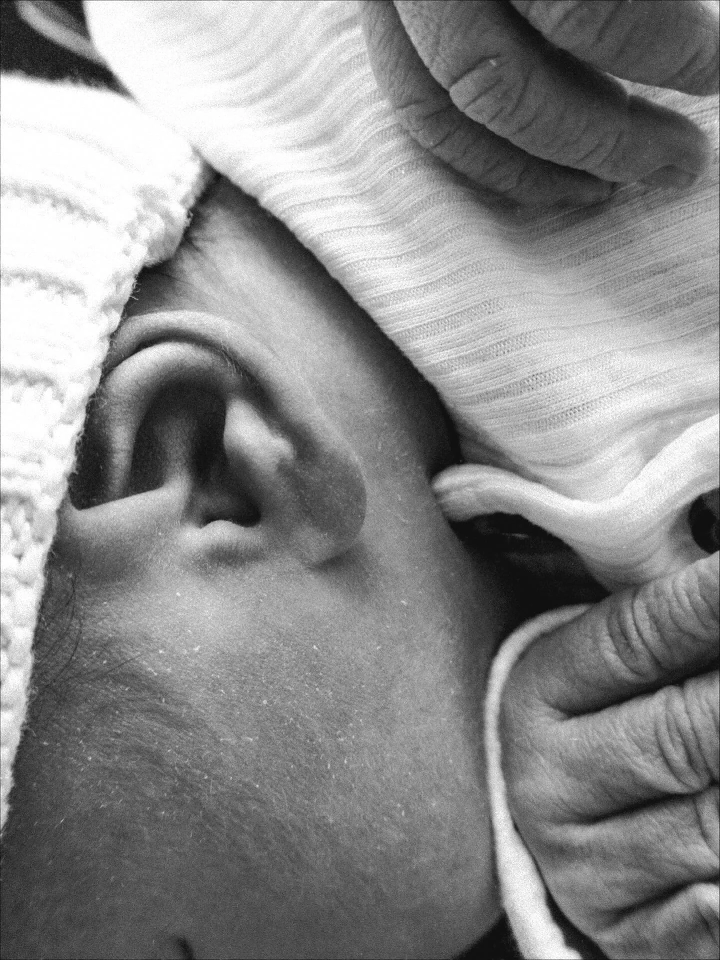 Close-up black and white photo of a baby sleeping, with part of an adult's hand gently holding the baby's head and head partially covered with a blanket.