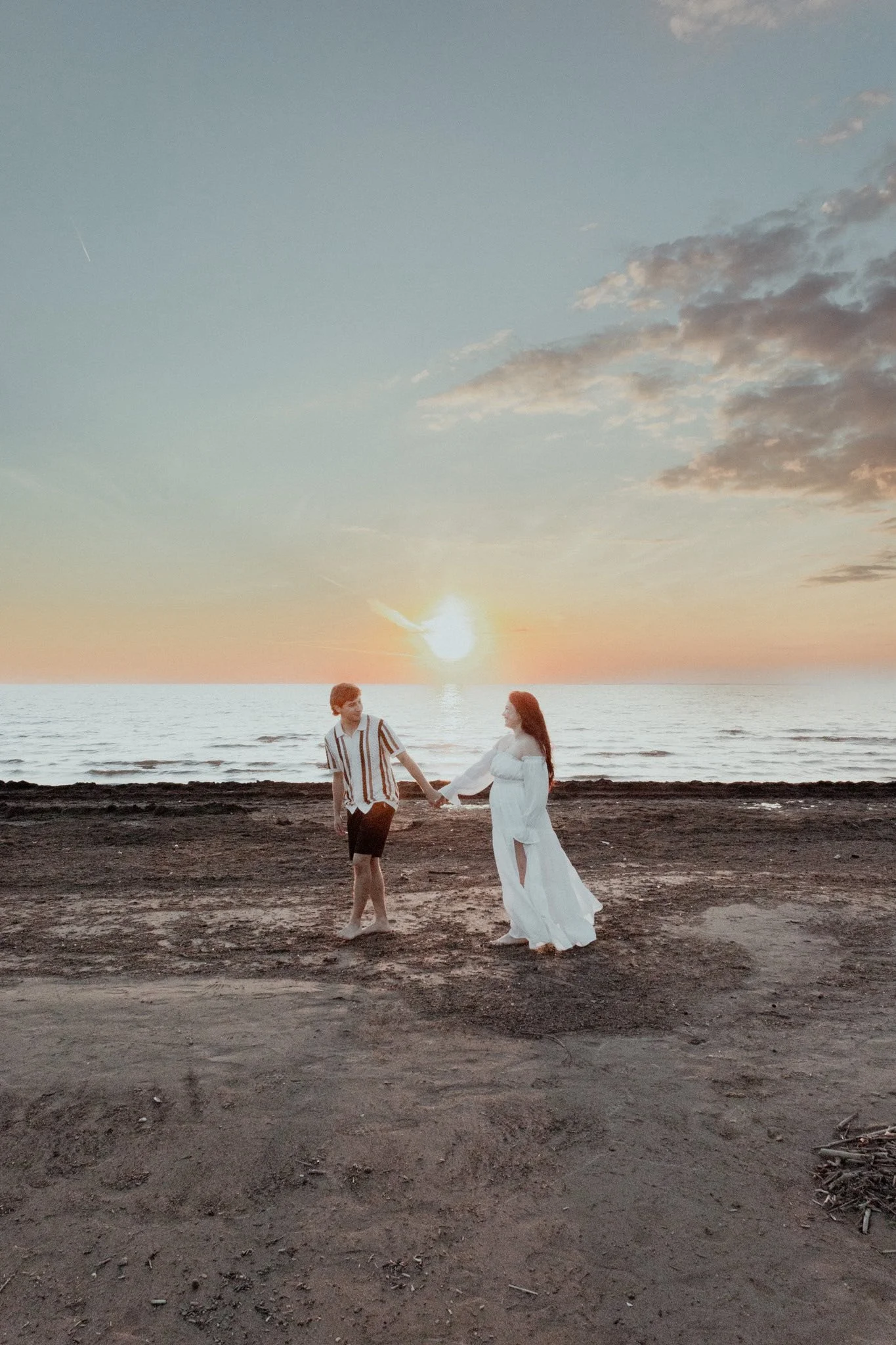 A couple holding hands on a beach at sunset, with the sun setting over the ocean in the background, the woman wearing a long white dress and the man wearing a striped shirt and shorts.