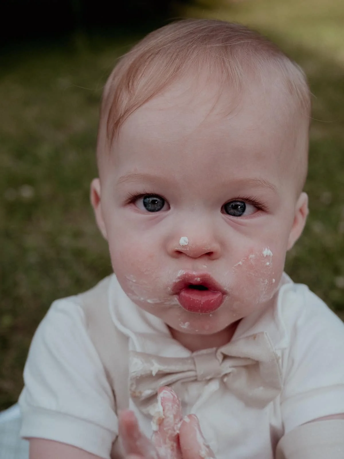 Close-up of a young child with blond hair, blue eyes, and a facial expression of surprise or curiosity, with food or cream on their face and hands, wearing a cream-colored shirt and bow tie outdoors grassy background.
