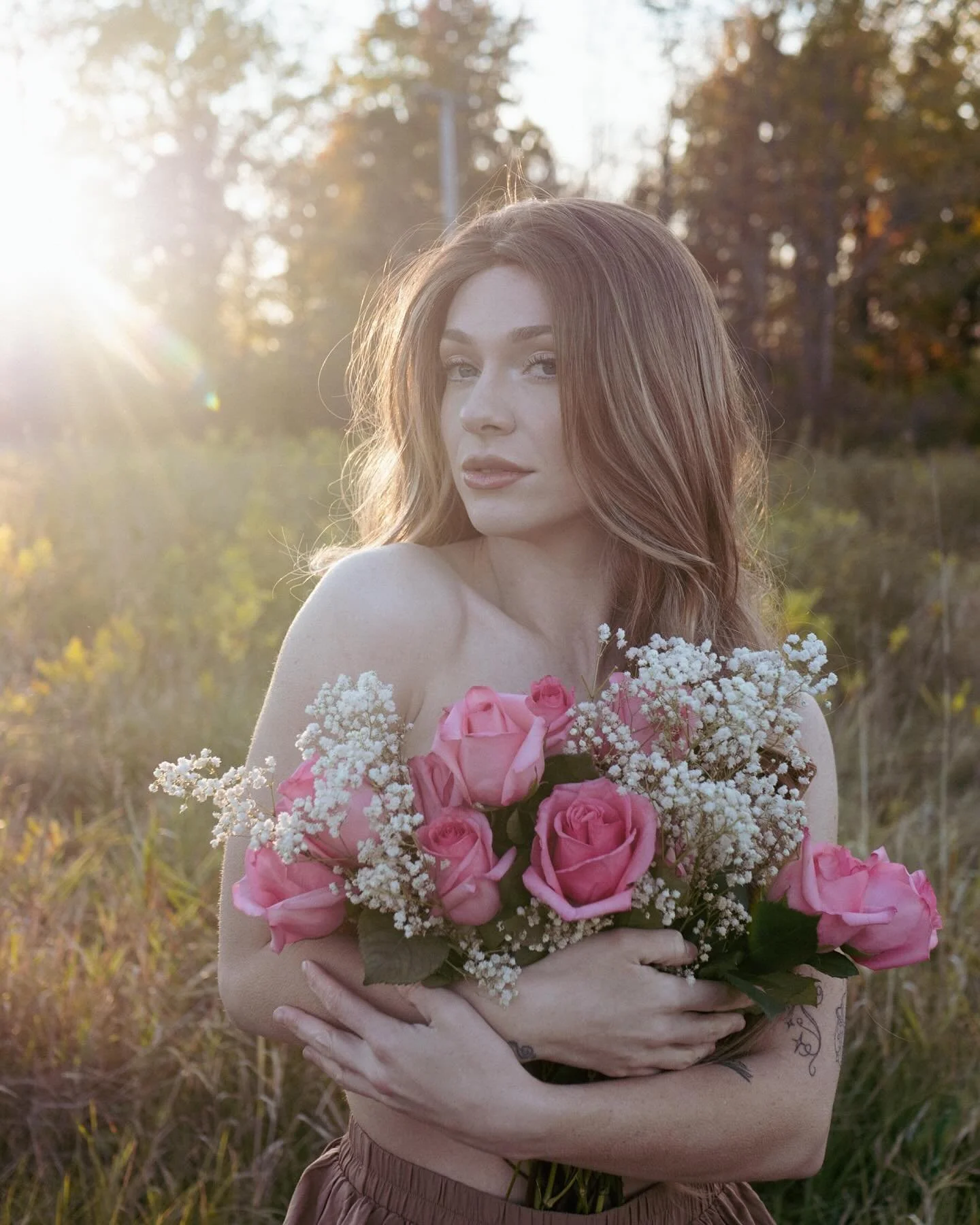 A young woman with red hair holding a bouquet of pink roses and white flowers outdoors during sunset