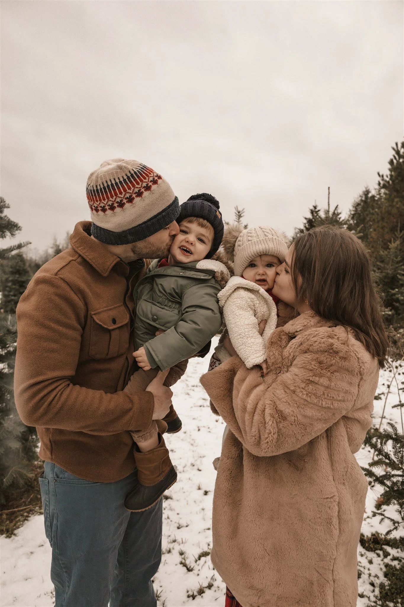 A family outdoors in a snowy christmas tree farm during winter, with the father and mother holding their two young children, all bundled in warm clothes and hats.
