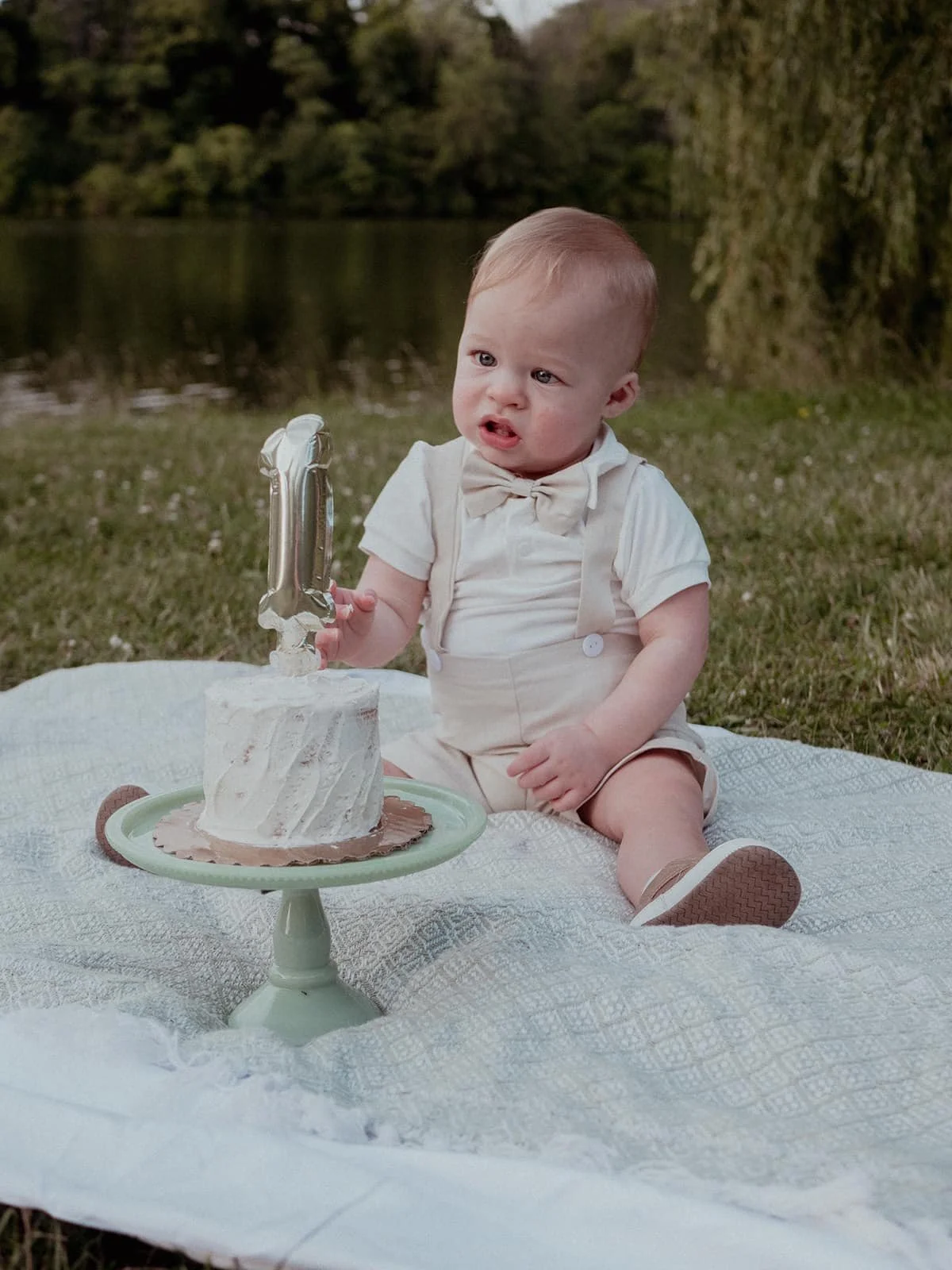 A young child sitting on a picnic blanket outdoors near a lake, holding a balloon shaped number 1, with a birthday cake in front of him.