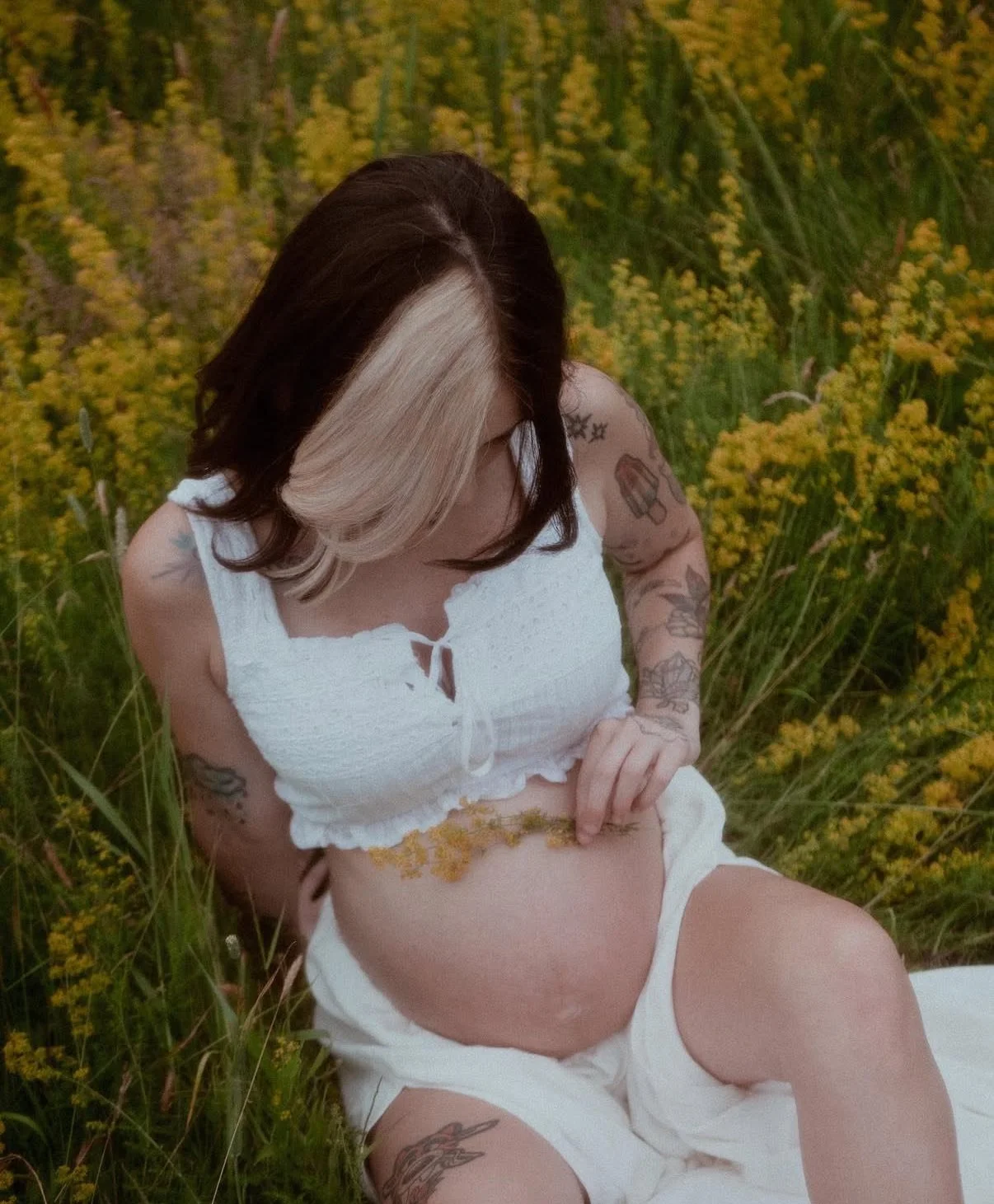 A pregnant woman with black and blonde hair, tattoos on her arms and leg, wearing a white crop top and shorts, sitting in a field of yellow flowers and looking at her belly.