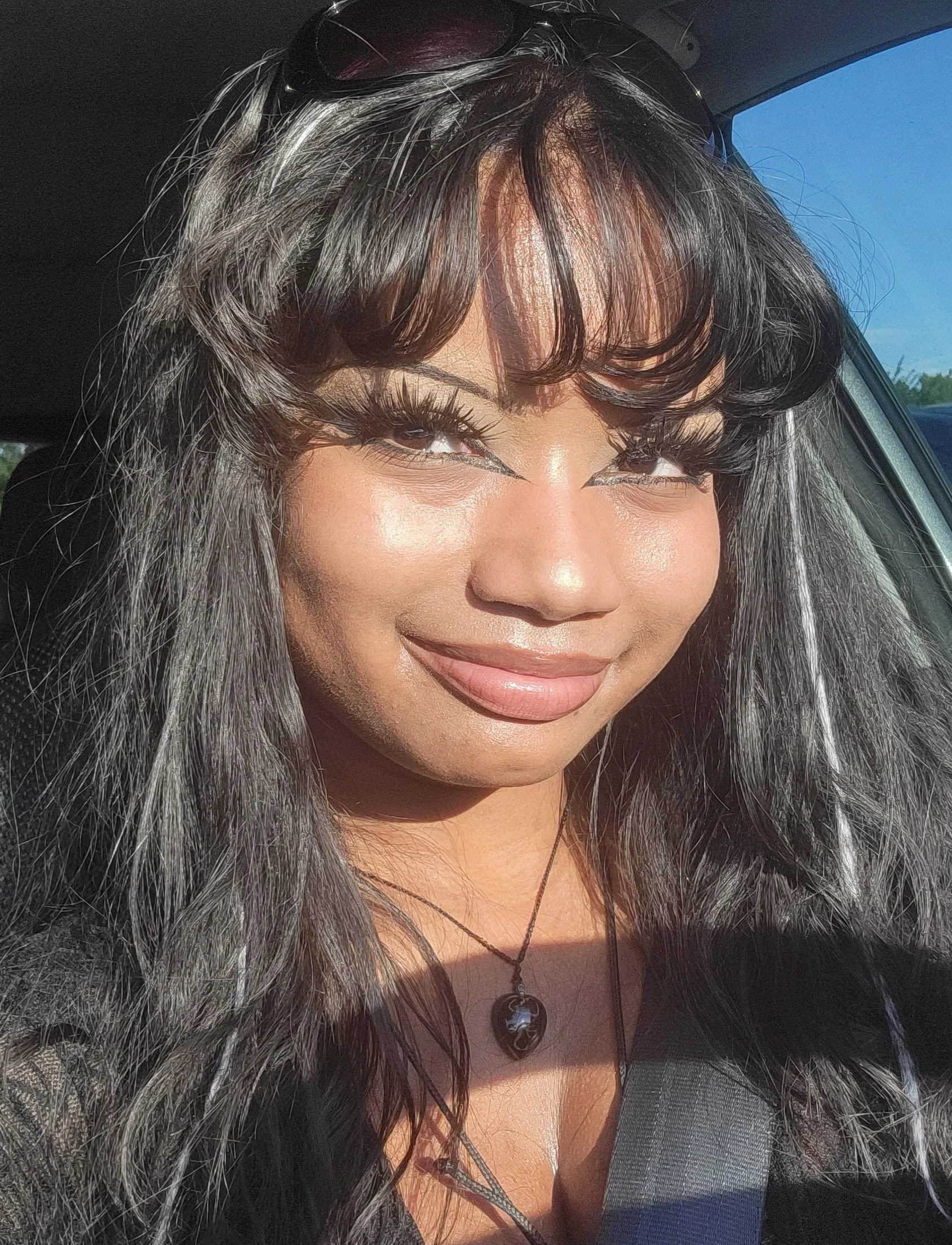 Close-up of a smiling woman with dark, wavy hair and bangs, wearing makeup with eyeliner and lipstick, black oval pendant necklace, sunglasses on her head, sitting in a car with sunlight on her face.