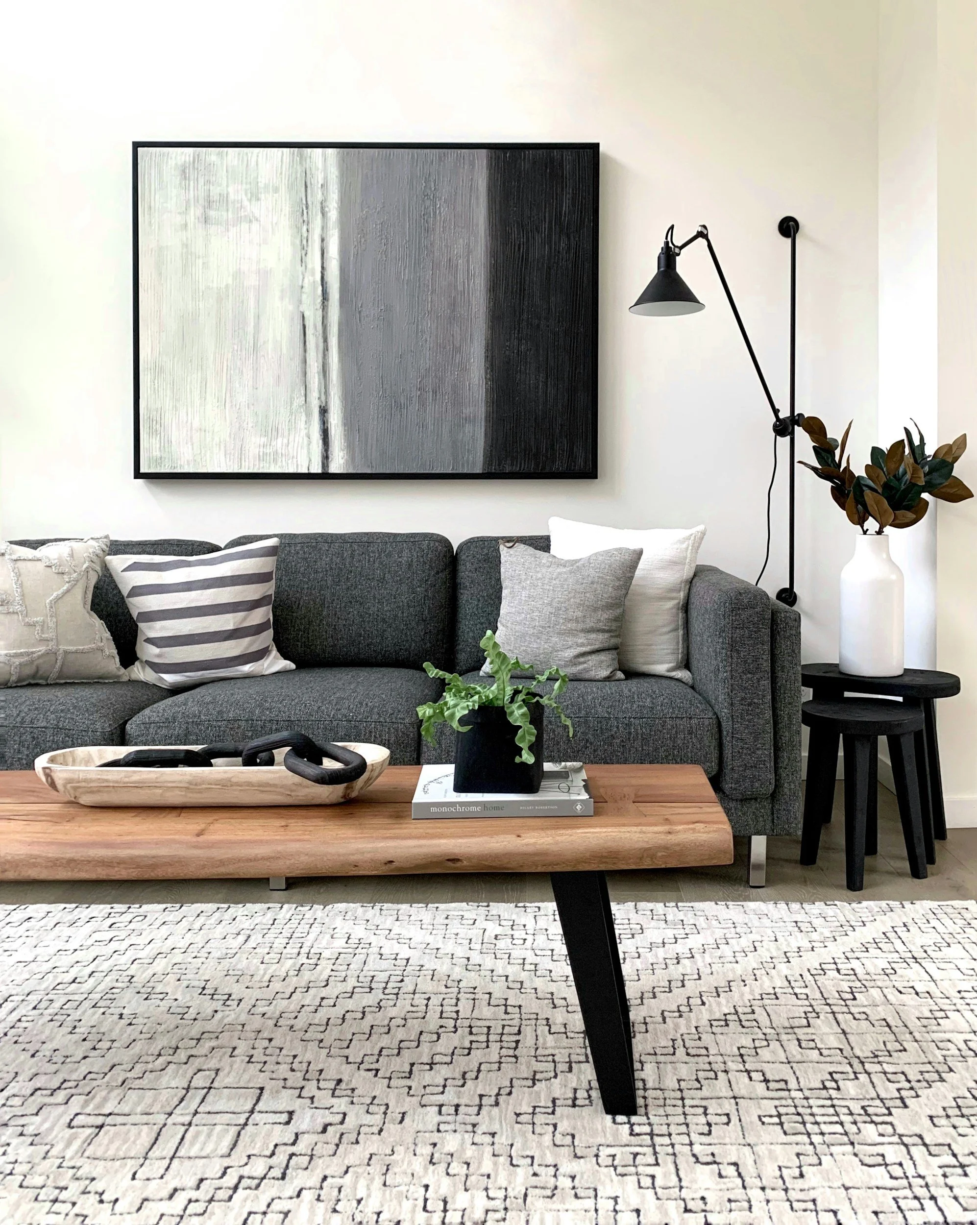 Modern living room with a dark gray sofa, black and white pillows, a wooden coffee table with a small plant and decorative black chains, a black side table with a white vase and plant, black wall-mounted lamp, abstract black, white, and gray artwork on white walls, and a light-colored patterned rug.