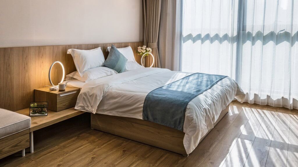 A neatly made bed with white sheets and pillows, a wooden headboard, and a blue throw blanket. There are modern table lamps, a flower vase, and a small photo frame on the bedside tables. A large window with sheer curtains lets in natural light, illuminating the wooden floor.