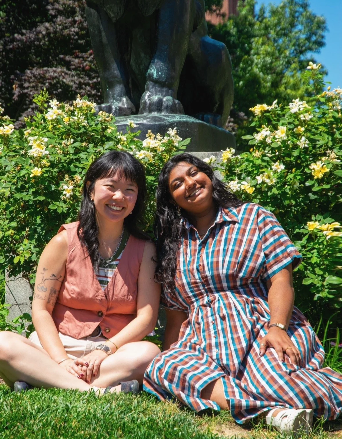 Two Queer and Asian Psycho-therapists smiling while sitting in the grass in front of a statue.