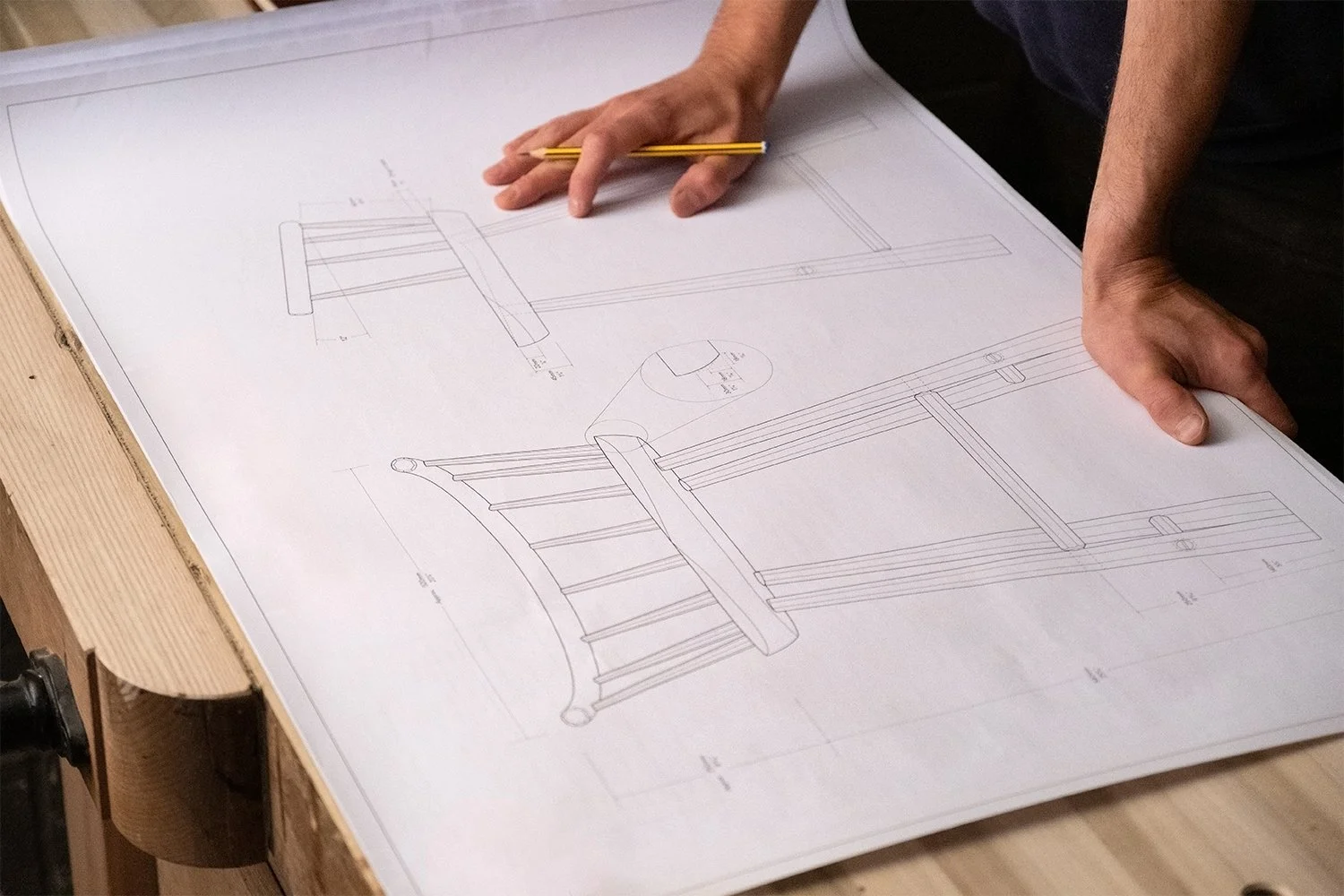 Person working on technical drawings or blueprints of furniture, possibly a chair, on a large sheet of paper on a work table.