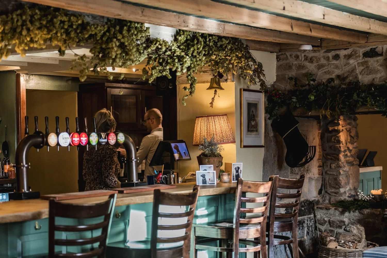Interior of a cozy, rustic bar or restaurant with a wooden bar counter, hanging beer taps, and a stone fireplace. There are framed photographs, a table lamp, and hanging floral decorations. Two people are chatting in the background near wooden cabine
