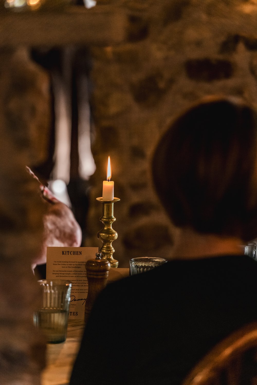 Dimly lit interior with a lit candle on a brass candlestick holder on a wooden table, surrounded by glasses and a small table sign labeled 'Kitchen,' with stone walls and a person in the background.
