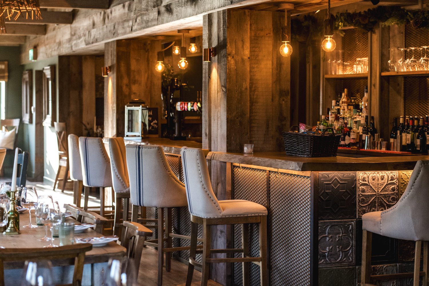 Interior of a cozy restaurant or bar with wooden decor, a bar counter lined with high-backed beige and striped chairs, and a shelf with various bottles of alcohol, glasses, and bar equipment, illuminated by warm hanging Edison bulbs.