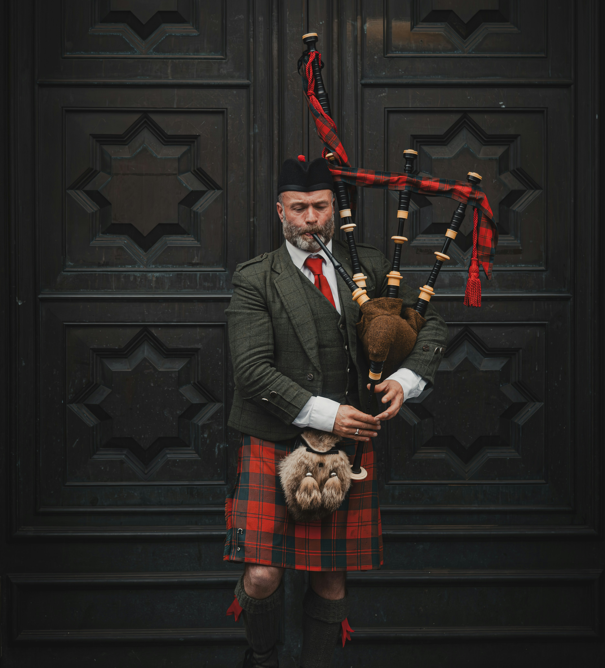 A man dressed in traditional Scottish attire playing bagpipes in front of a black wooden door  ready for a burns night celebration.