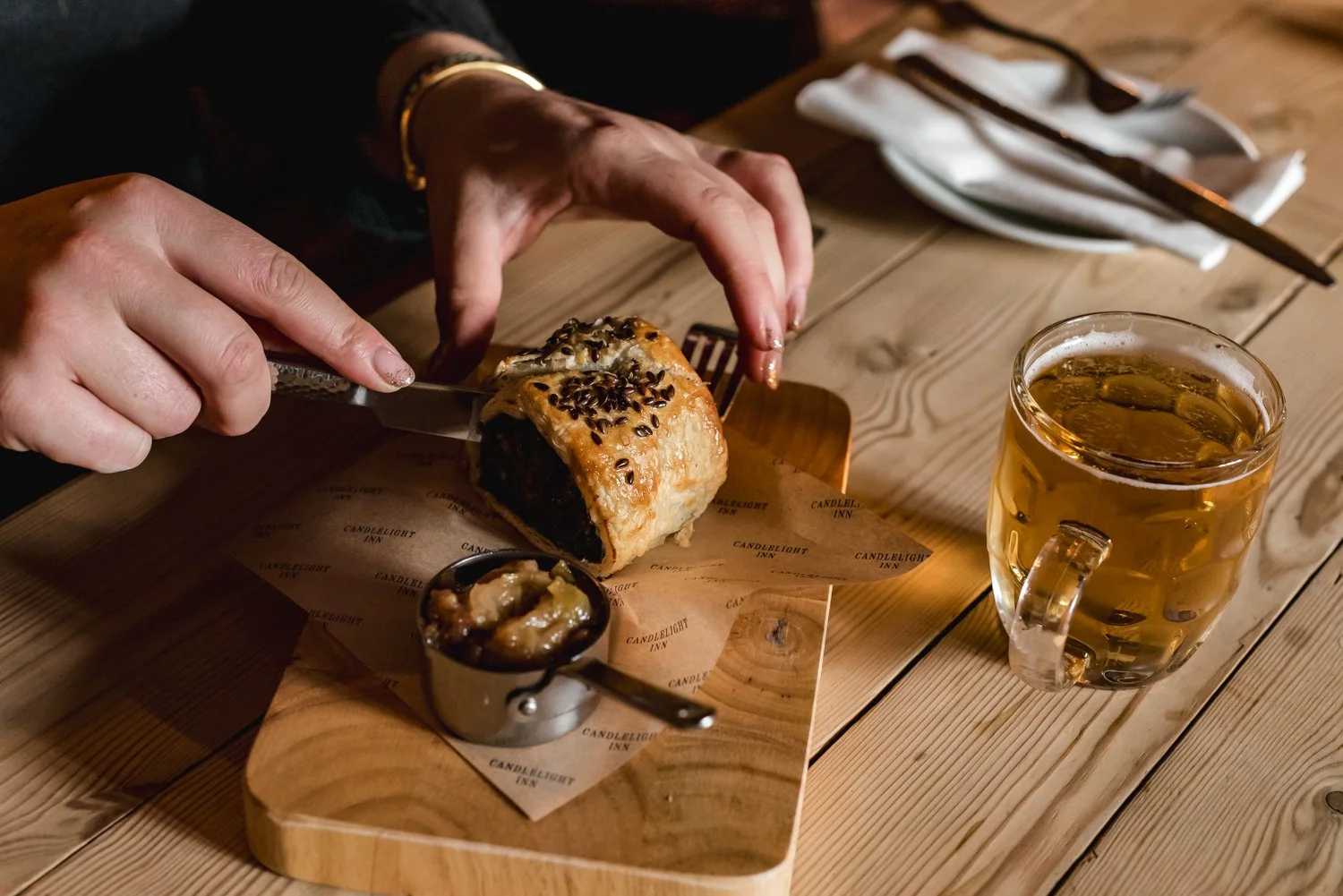 Person slicing a pastry topped with chocolate shavings on a wooden board, with a small dish of caramelized fruit, a glass of beer, a white napkin with cutlery, and a fork on a wooden table.