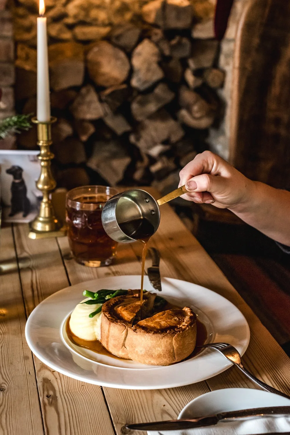 A person pouring gravy over a Yorkshire pudding on a white plate, accompanied by green beans and mashed potatoes, in a rustic dining setting with a lit candle and a glass of tea.