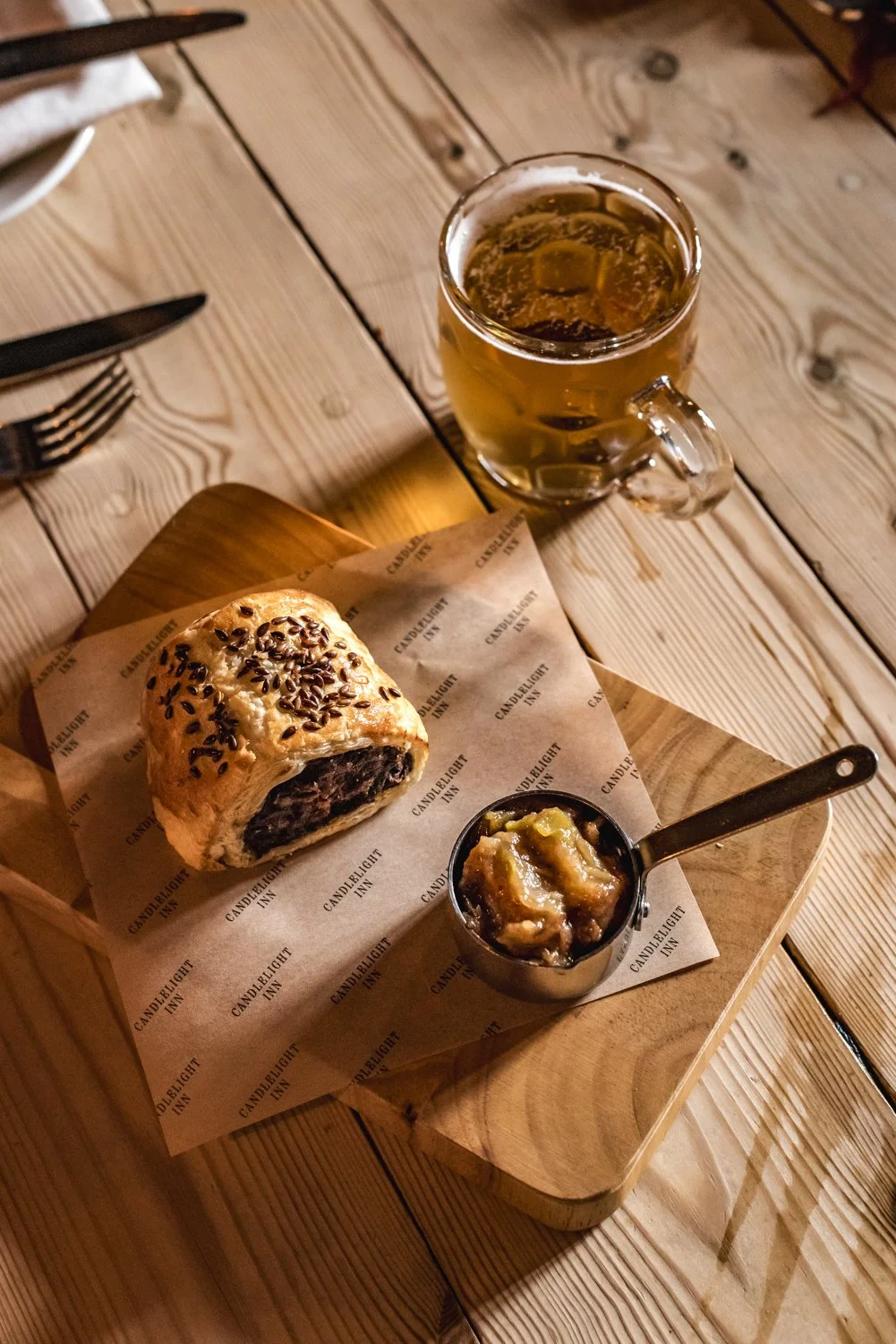 A cherry Danish pastry with chocolate sprinkles, a small container of apple butter, a glass mug of beer, and a set of black utensils on a wooden table.