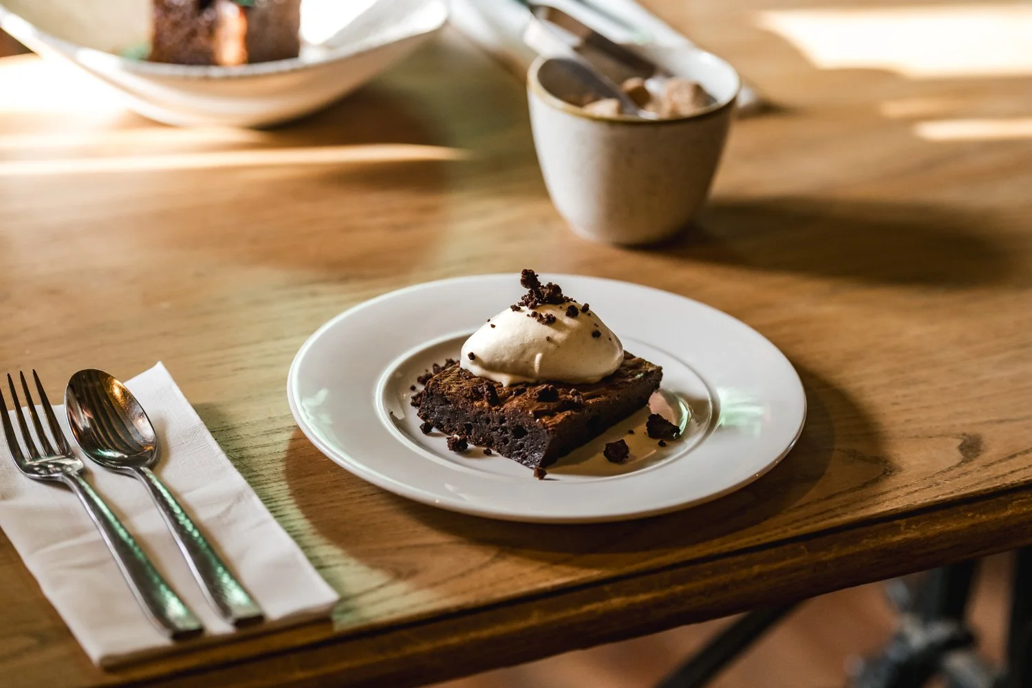 A slice of chocolate brownie topped with vanilla ice cream and chocolate crumbs on a white plate, with a fork and spoon on a napkin to the left, on a wooden table, with a bowl and spoon in the background.