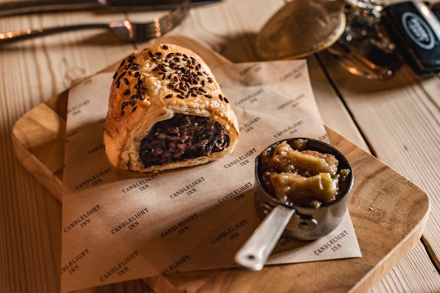 A pastry with a flaky crust and chocolate filling, topped with sesame seeds, placed on parchment paper with 'CANDLELIGHT INN' printed repeatedly, alongside a small metal cup of jam or preserve on a wooden tray.