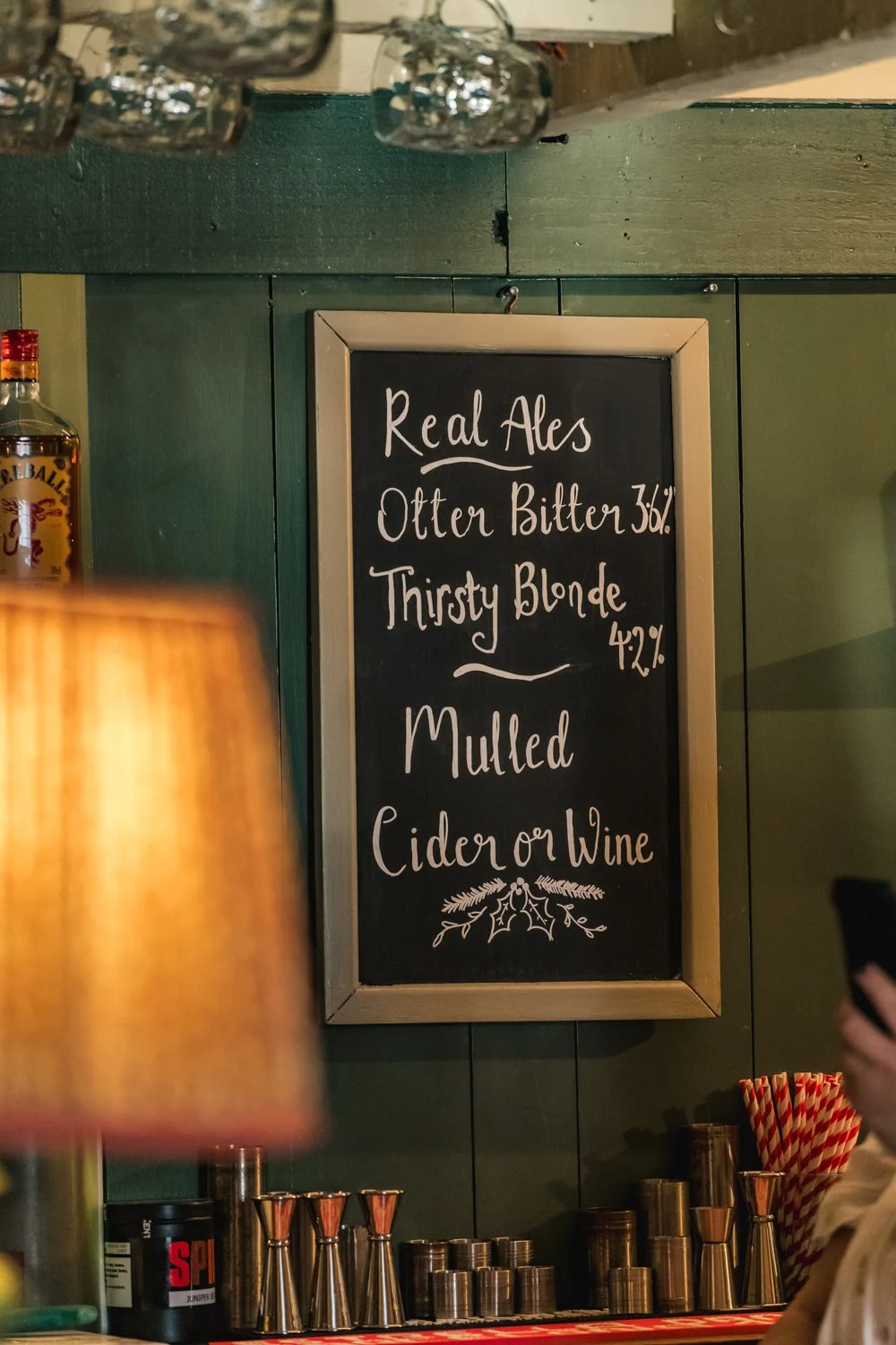 A chalkboard menu in a bar listing craft beers and their alcohol content, with a bottle of alcohol and bar tools visible nearby.