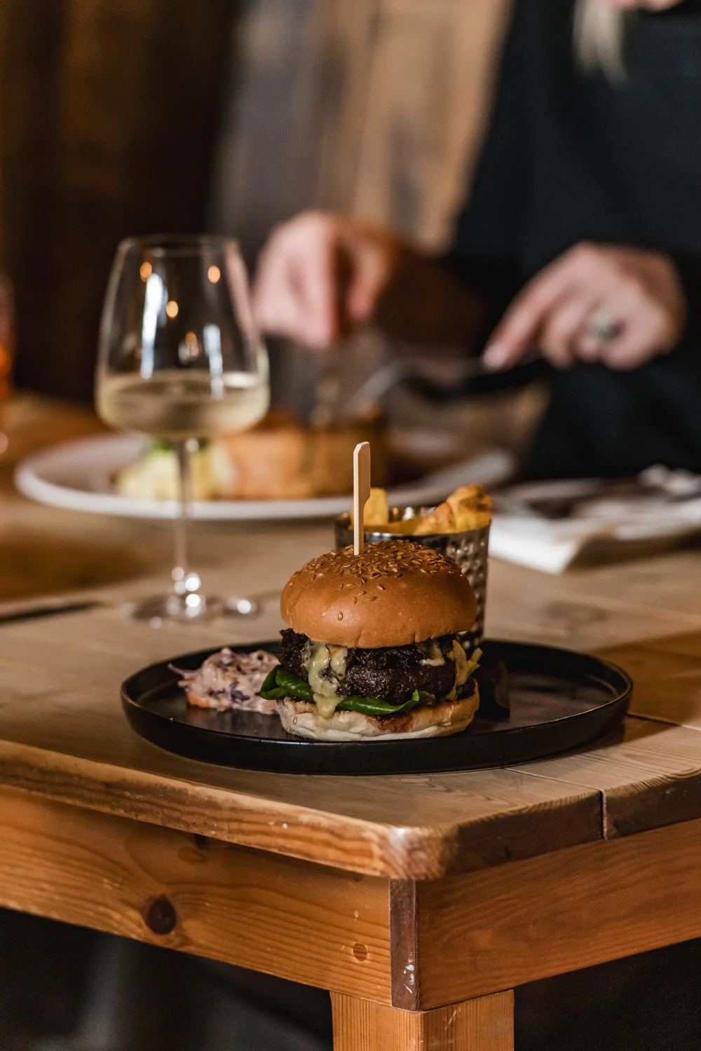 A gourmet burger with a beef patty, melted cheese, spinach, and sauce in a bun with sesame seeds, served on a black plate with a toothpick, accompanied by fries in a metal cup, on a wooden table with a glass of white wine in the background.
