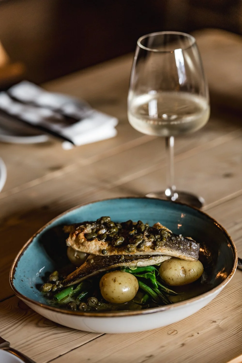 A plate with fish topped with capers, accompanied by green beans and denver potatoes, on a wooden table, with a glass of white wine in the background.