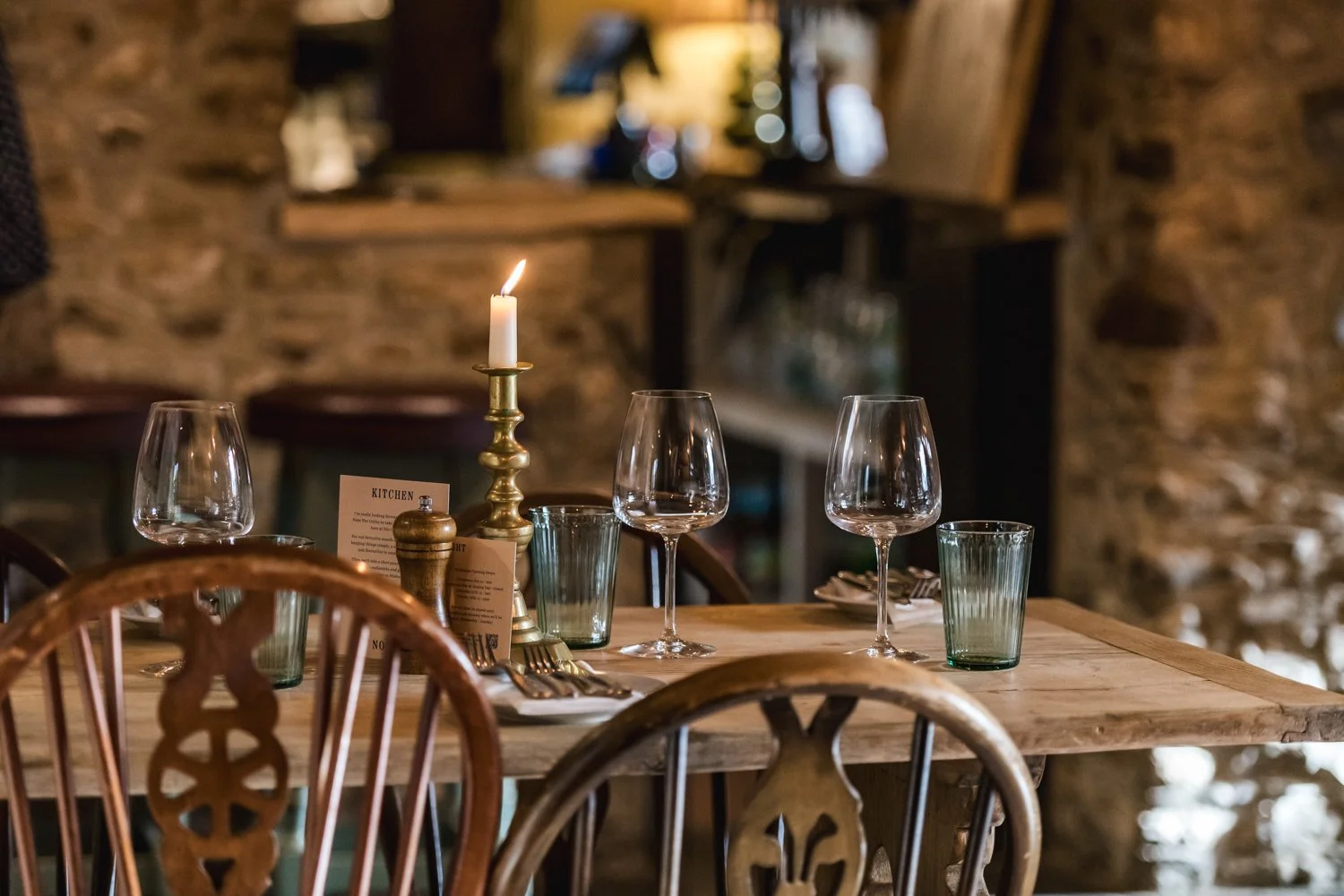A rustic dining table set with three glasses of water, two empty wine glasses, a lit candle in a vintage brass candlestick, and a small menu card in a cosy restaurant with exposed stone walls.