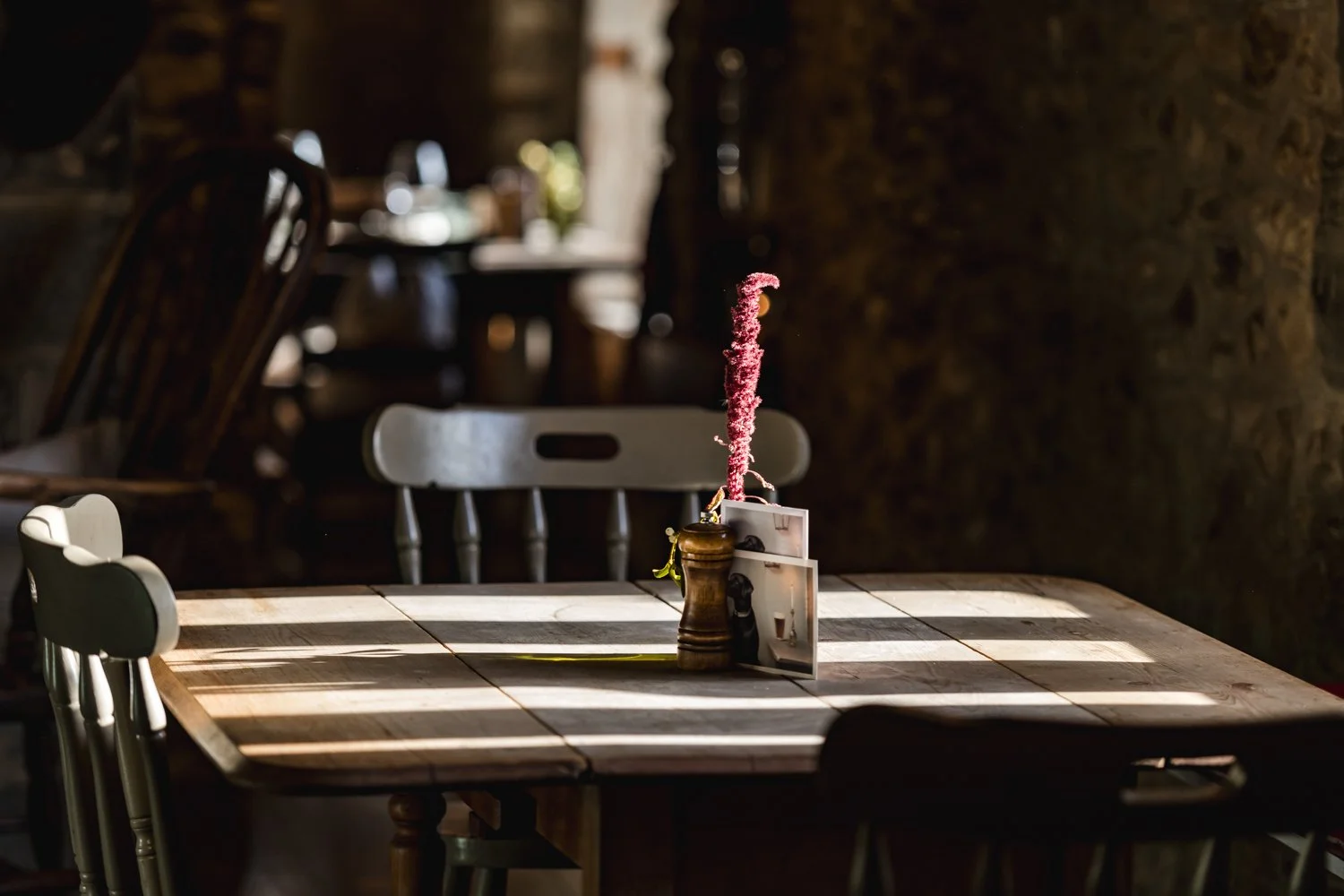 A wooden table with a small vase holding a pink sparkler, surrounded by photographs and a pepper shaker, illuminated by sunlight in a cozy cafe.