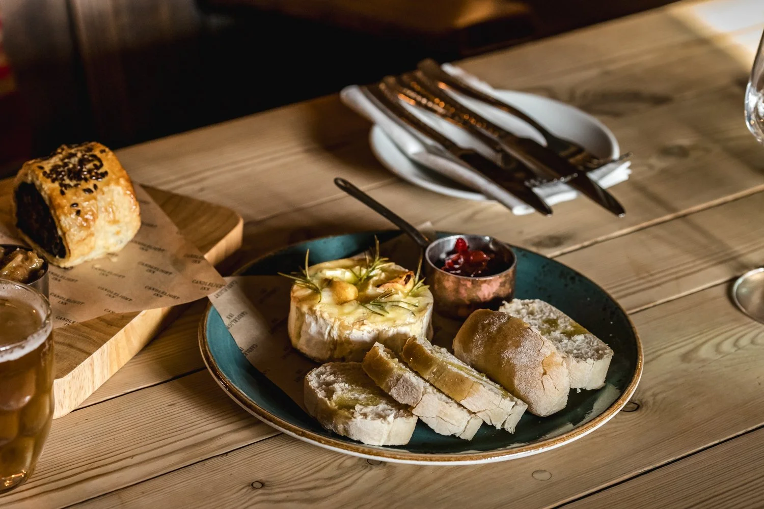 A plate with bread, cheese, and jam on a wooden table, along with a pastry, cutlery, and a glass of beer.