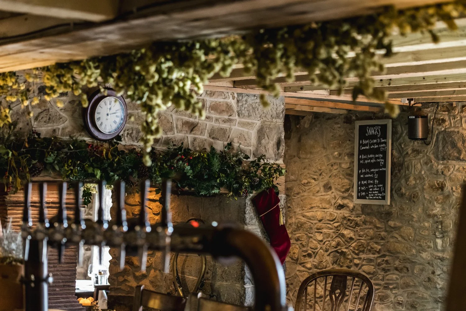Interior of a rustic restaurant with stone walls, a wall-mounted clock, hanging dried herbs or flowers, and a black chalkboard menu listing snacks, decorated for the holidays with a red stocking hanging on the wall.