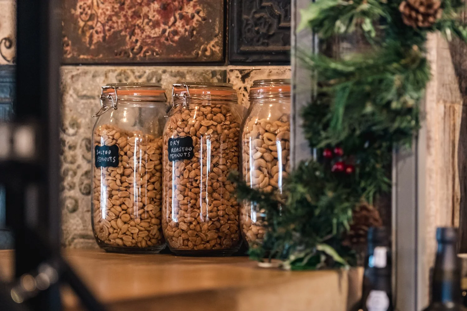 Glass jars filled with salted peanuts, dry roasted peanuts, and another type of peanuts on a wooden shelf in a rustic kitchen, with a decorative plant in the foreground.