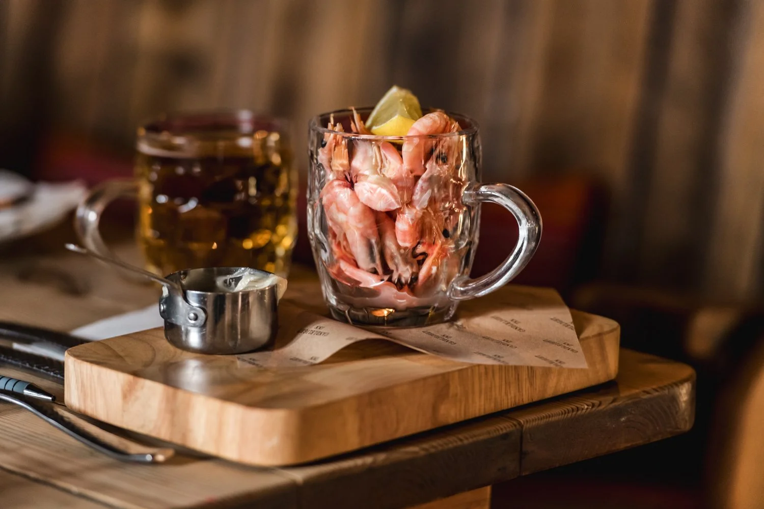 Clear glass mug filled with cooked pink shrimp and lemon wedge on top, on a wooden cutting board with a small metal container of cocktail sauce nearby, in a cozy restaurant setting.