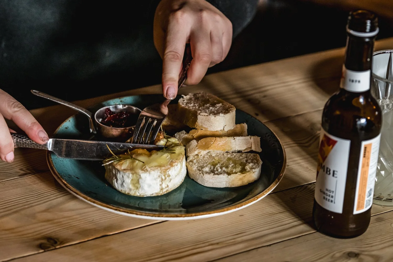Plate of sliced bread, soft cheese, and a small bowl of jam with a person cutting into the cheese with a fork and knife, on a wooden table with a bottle of beer nearby.