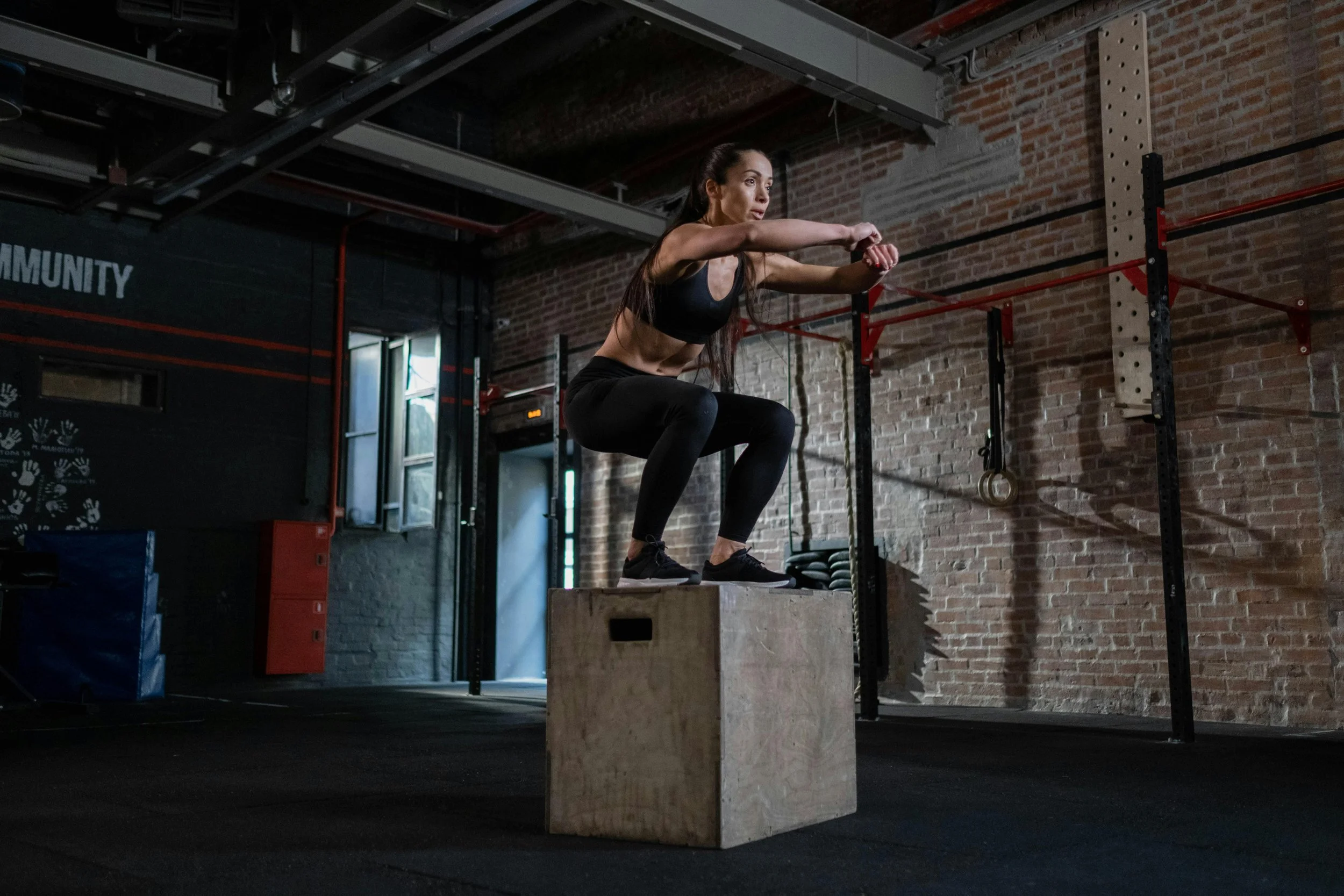A woman in workout clothes is performing a squat exercise on a wooden box in a gym with exposed brick walls and fitness equipment.