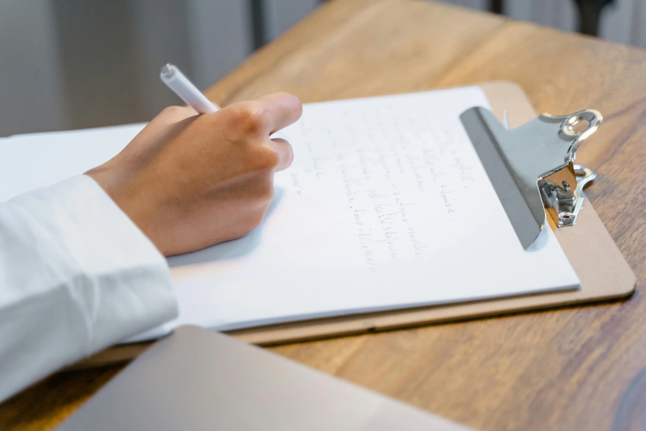 A person with light skin and wearing a white long sleeve shirt writing on a piece of paper clipped to a clipboard on a wooden table.