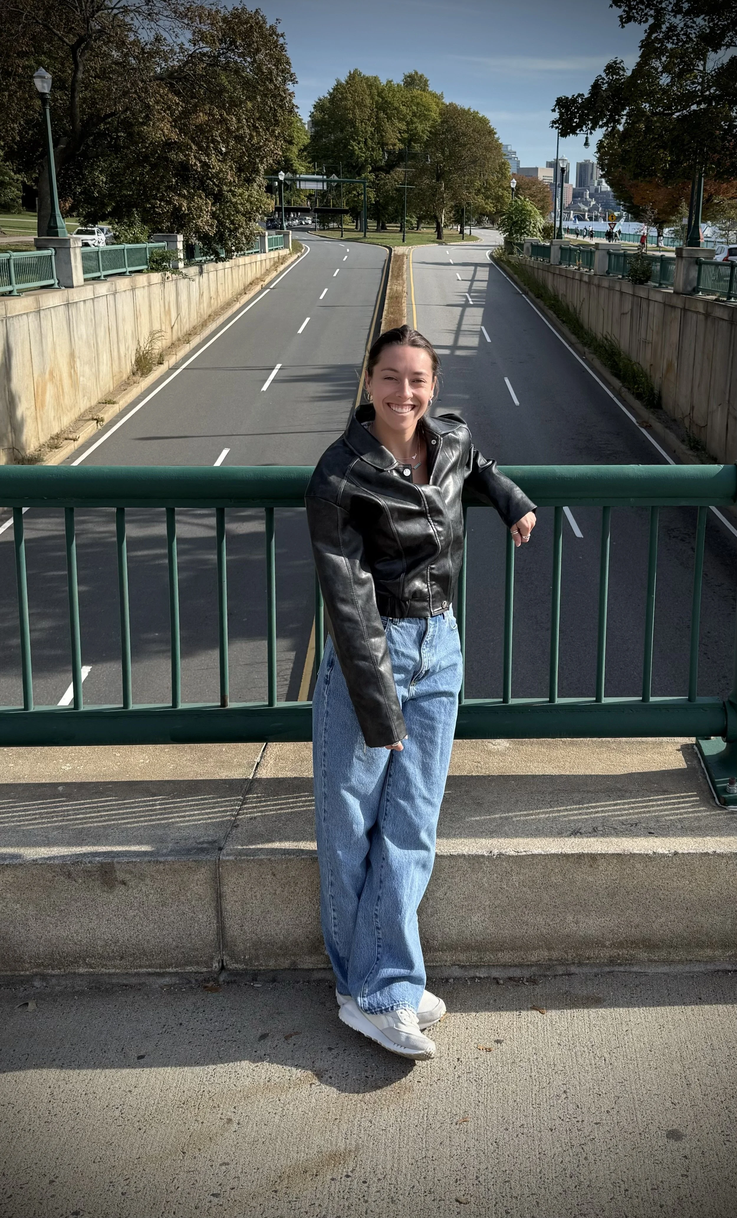 A young woman in a black leather jacket and wide-leg jeans stands on a bridge with green railings, smiling at the camera. Behind her is a cityscape with trees, a road, and buildings in the distance on a sunny day.