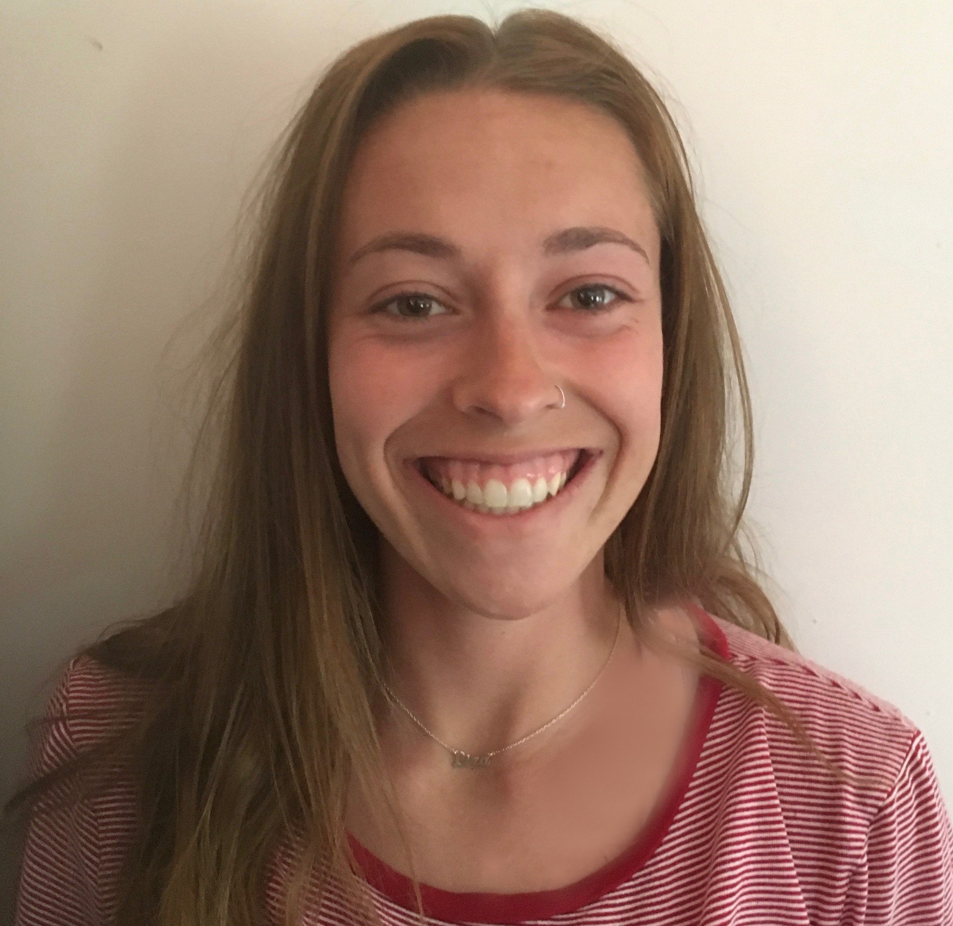 A woman with long light brown hair, wearing a red and white striped shirt, smiling, standing against a plain light-colored wall.