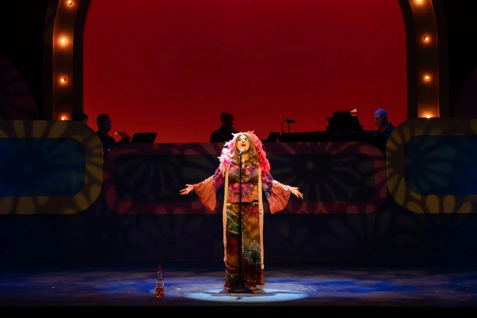 Ana Kusenberger playing Janis Joplin, standing center stage surrounded by flashbulb lights, arms extended outwards.