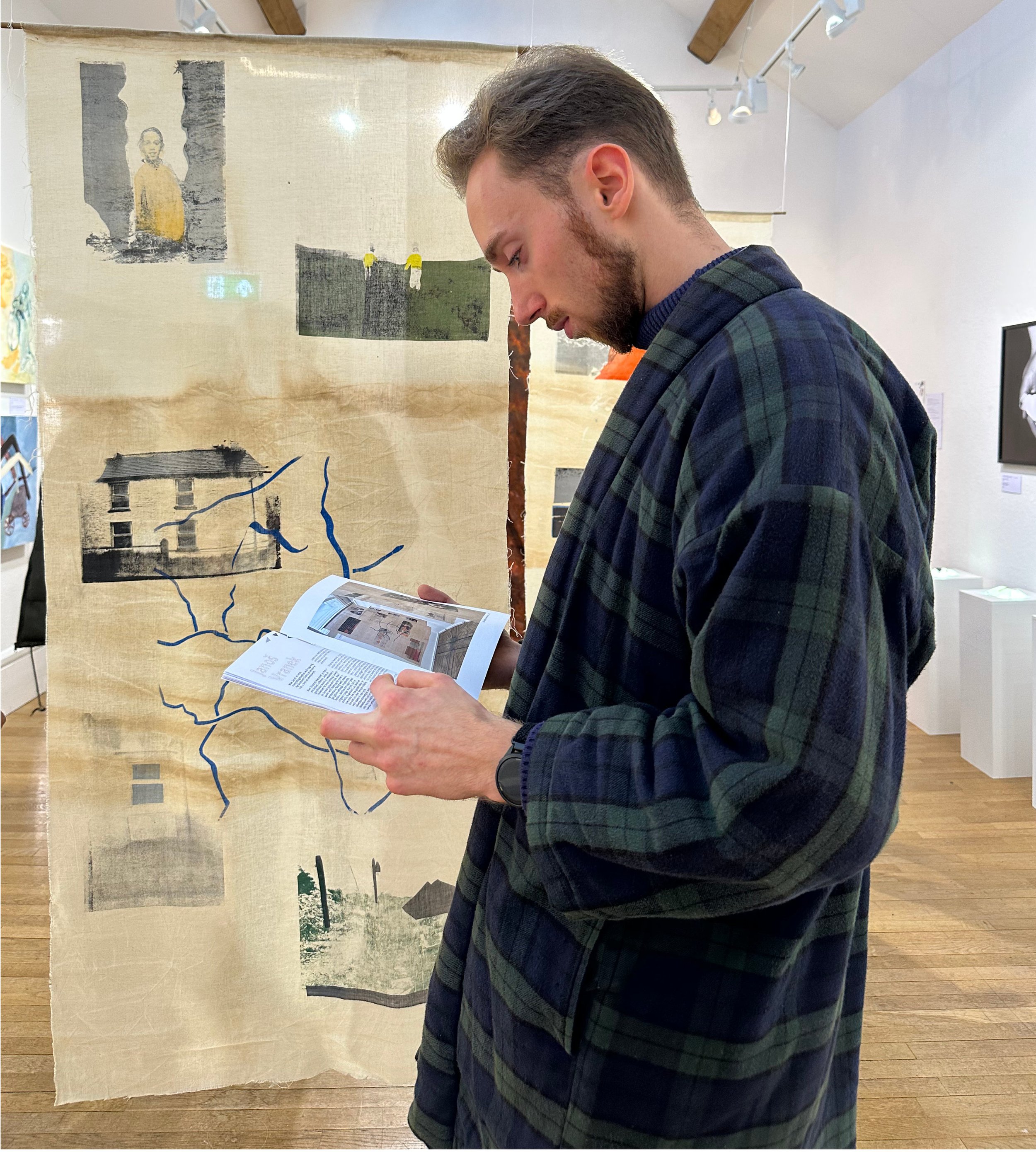 A young man side on, in a green and blue jacket reading a booklet in an art gallery with hanging textile art in the background.