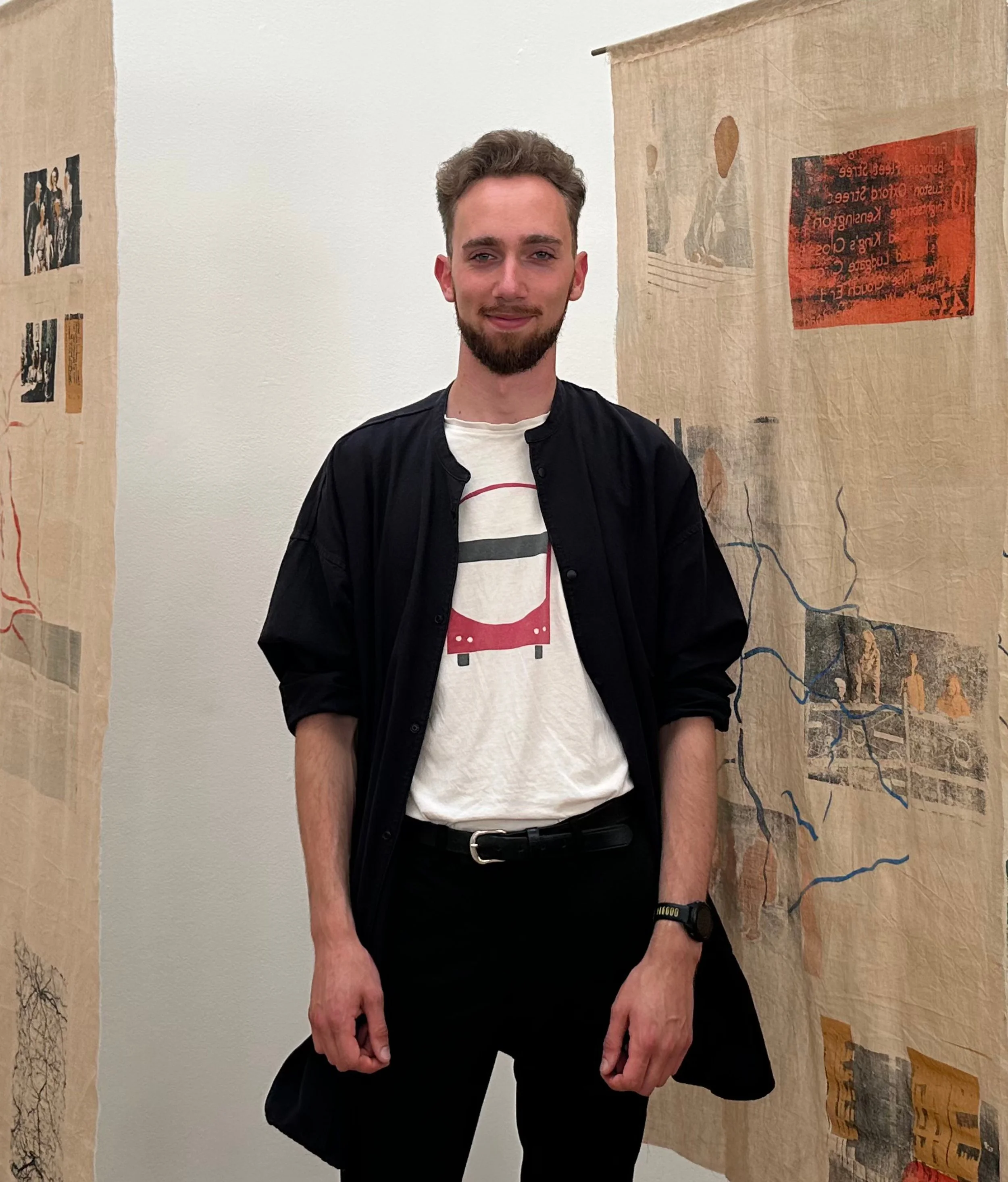 A young man stands in an art exhibit, wearing a black jacket over a white T-shirt with a screen-printed motif, black trousers, and a black watch. Behind him are collaged printed artworks on beige fabric hangings.