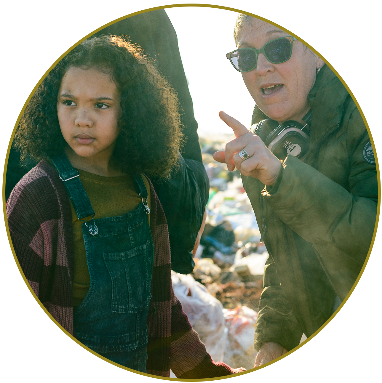 A woman wearing sunglasses and a green jacket points and talks to a young girl with curly hair, wearing a plaid sweater and denim overalls, near a pile of trash outdoors.