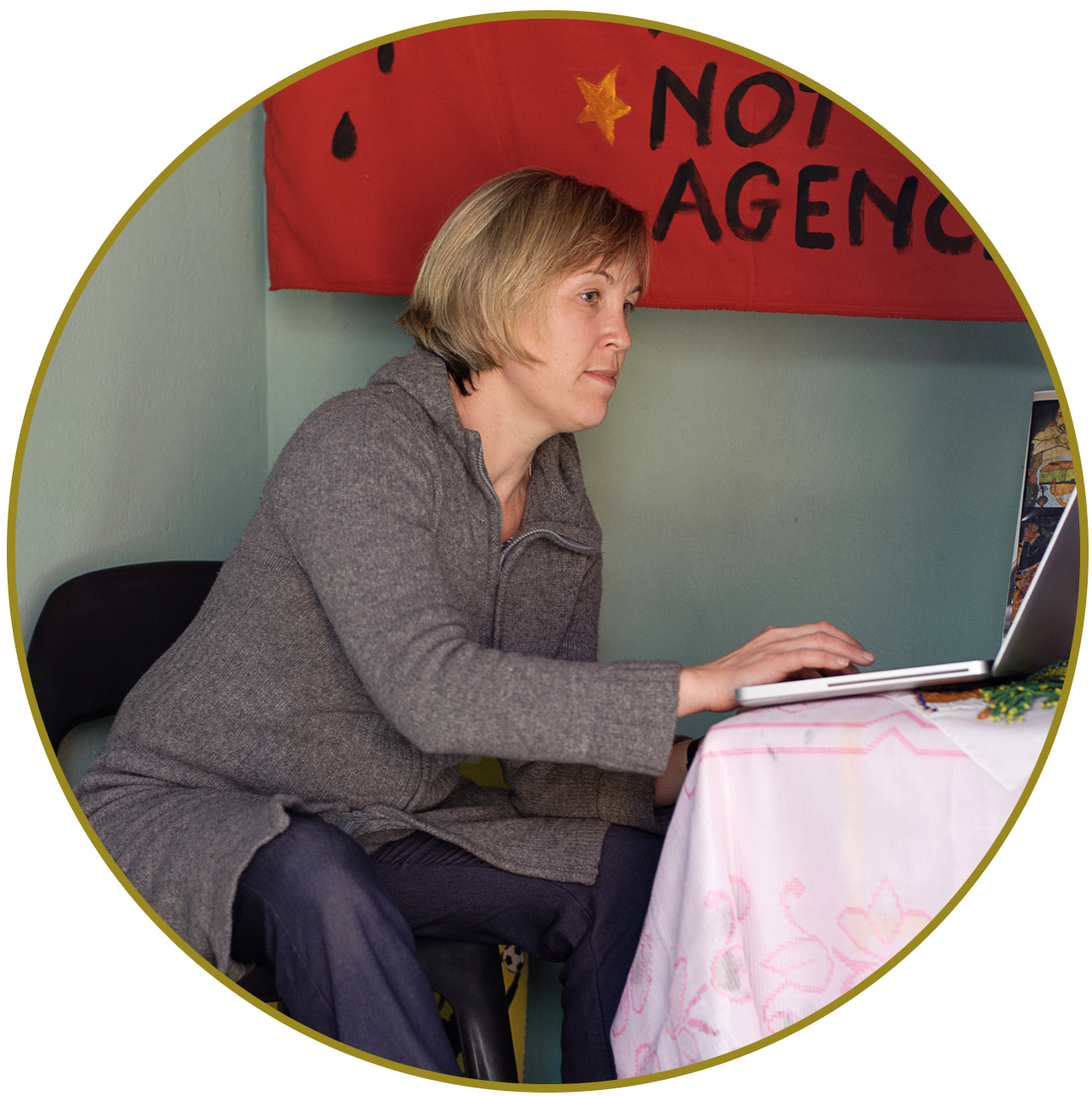 A woman with short blonde hair sitting at a table, working on a laptop. Behind her is a red tapestry with black lettering that says 'NOT AN AGENCY' and decorative stars.