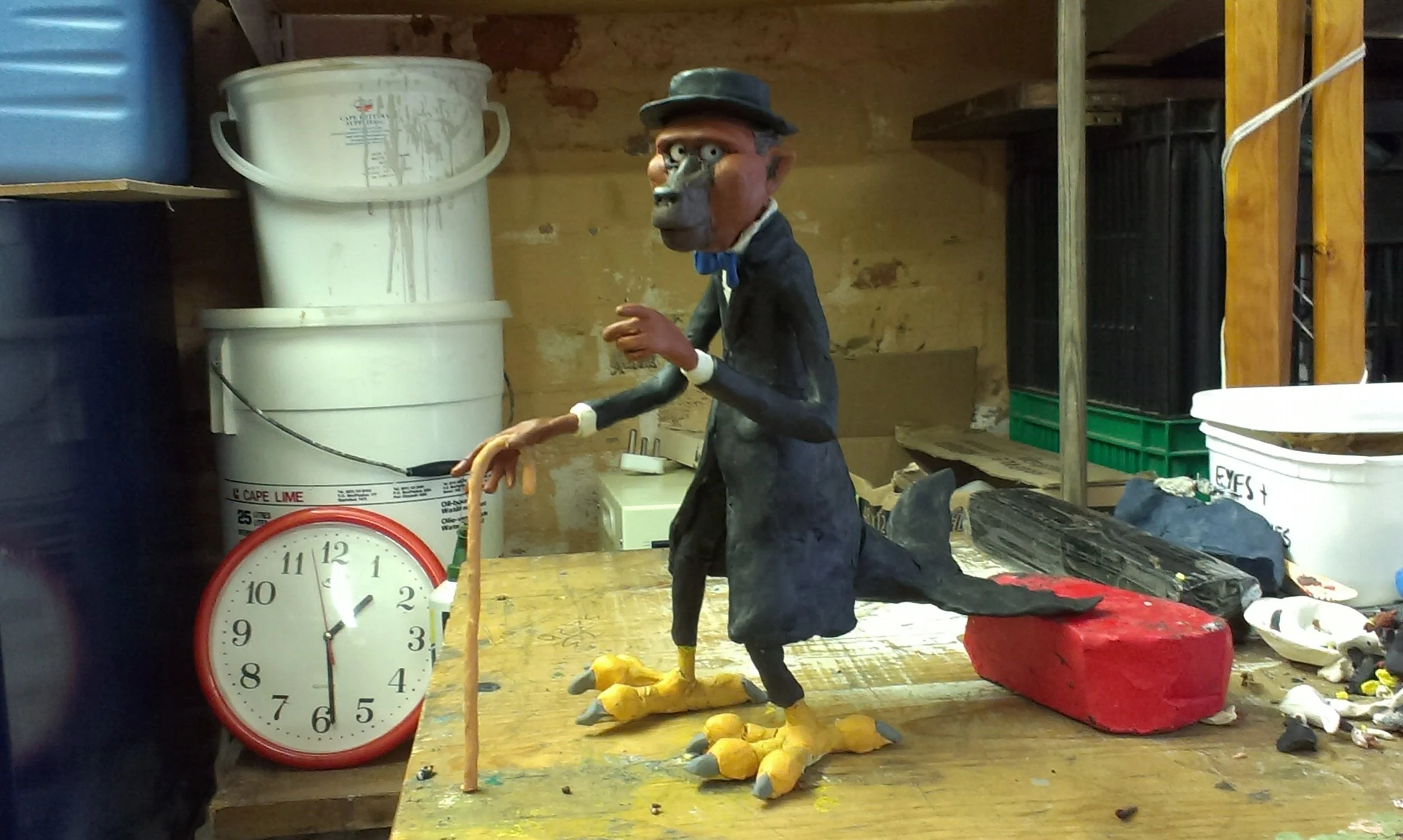 A detailed sculpture of an old man with a long grey beard wearing a hat, suit, and bow tie, standing on a wooden table with bird-like feet, holding a cane, in a workshop surrounded by buckets, tools, and craft supplies.