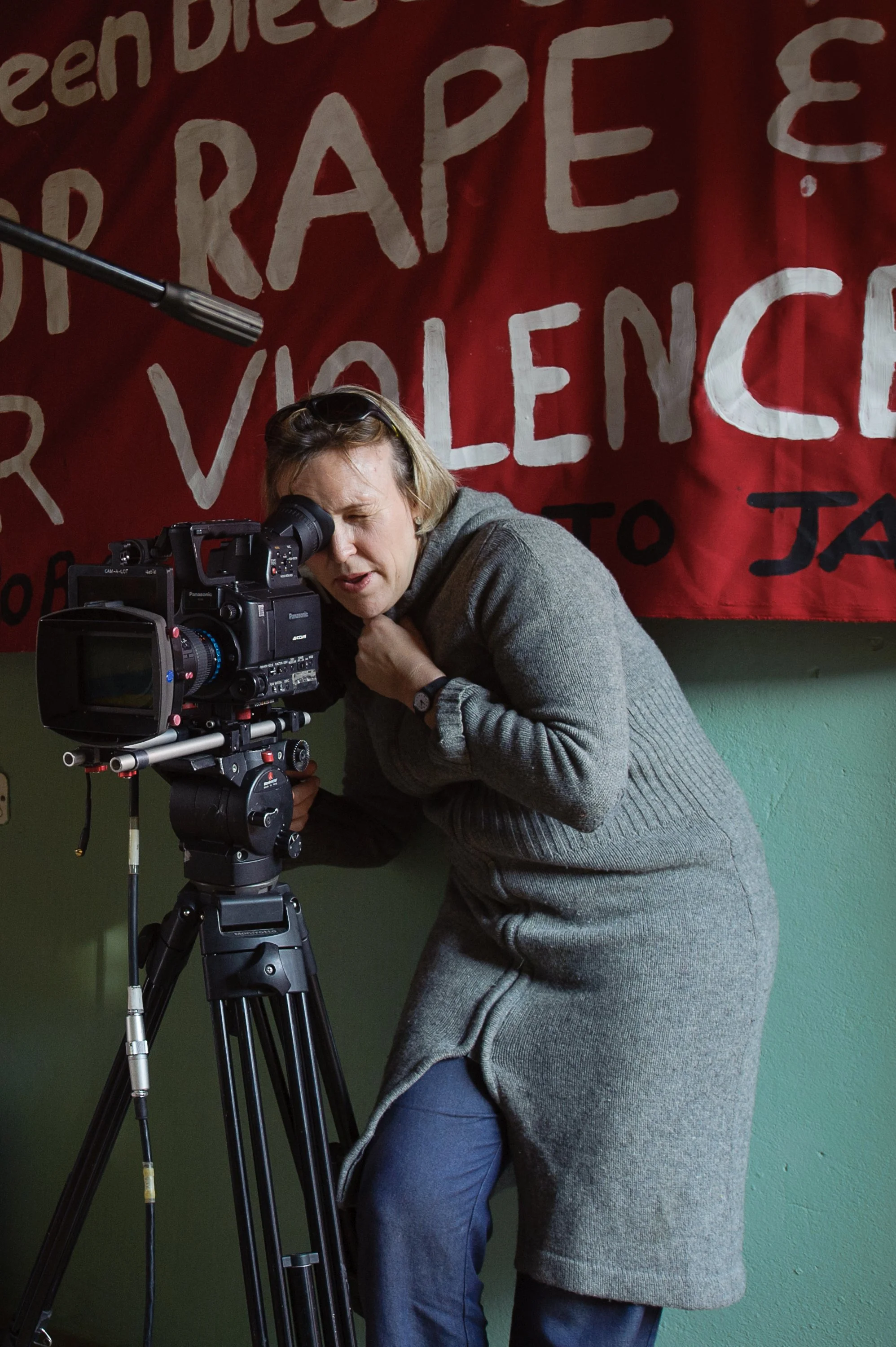 A woman operating a professional video camera on a tripod, looking into the viewfinder, in front of a red banner with political slogans.